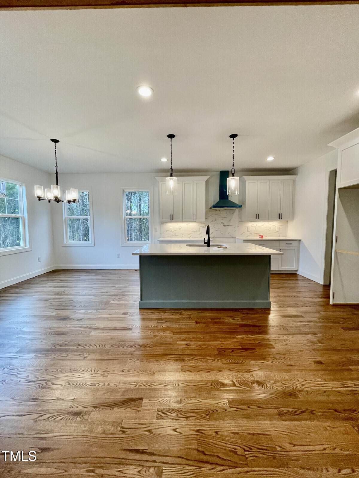 Modern kitchen with light wood floors, white cabinets, center island with sink, two pendant lights, three tall windows, and a chandelier over the dining area. Marble backsplash and teal range hood add contrast.