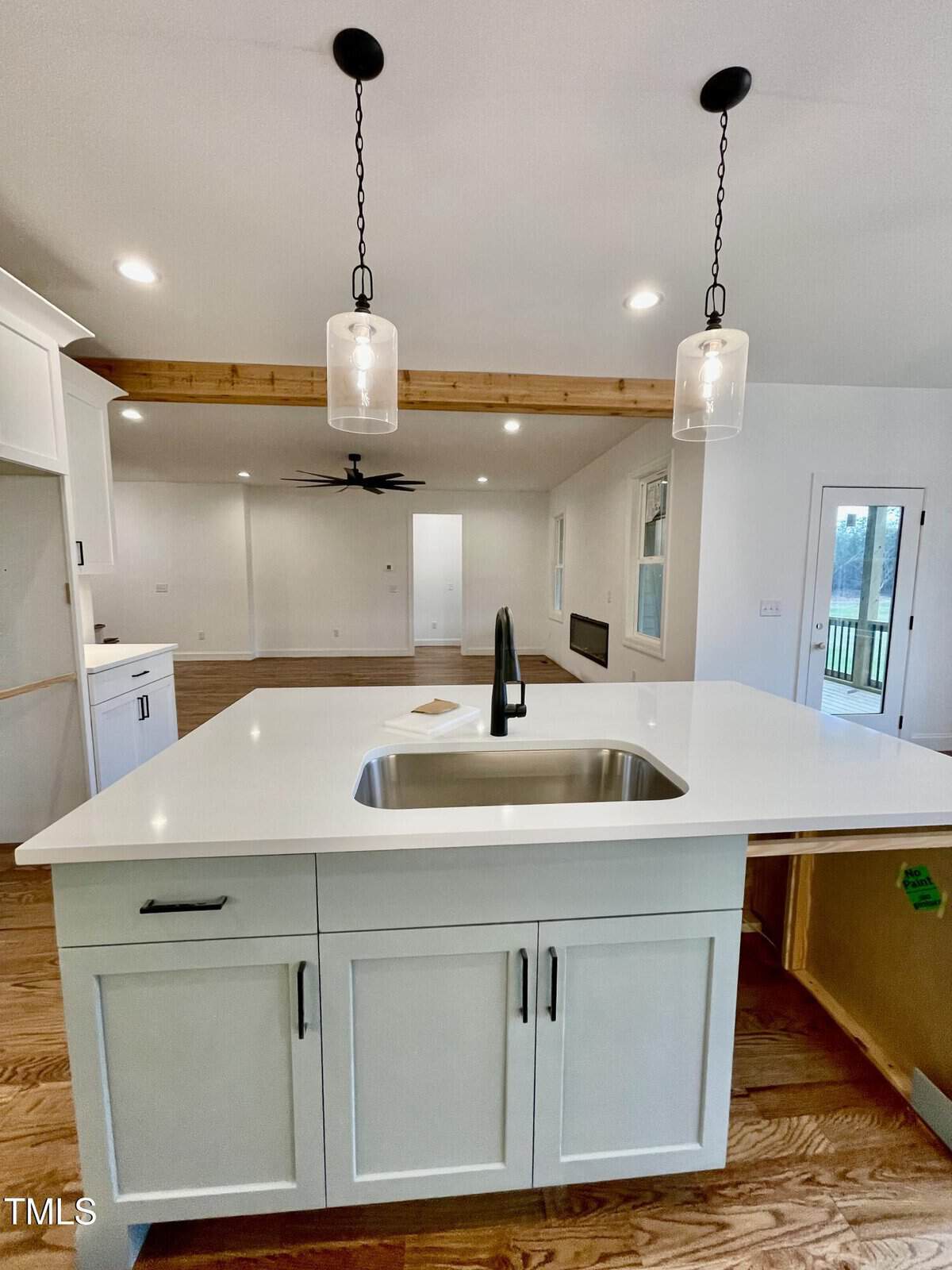 Bright modern kitchen with a white island featuring a sink and black faucet, pendant lights above, wood floors, exposed ceiling beam, and an open living area with large windows in the background.