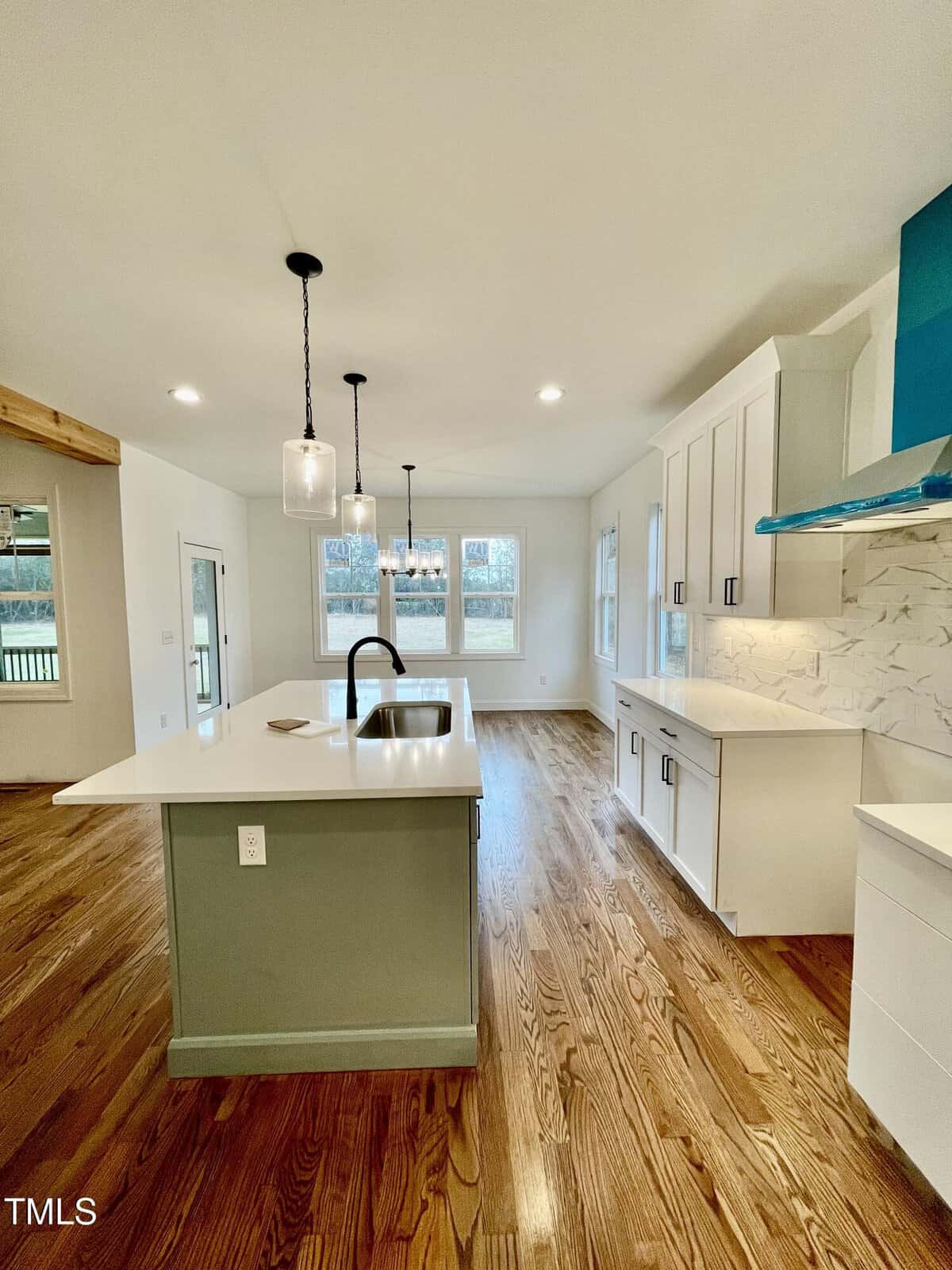 Bright, modern kitchen with white cabinets, marble backsplash, a green island with sink, pendant lights, and hardwood floors. Large windows let in natural light, and open shelving is visible on the wall.