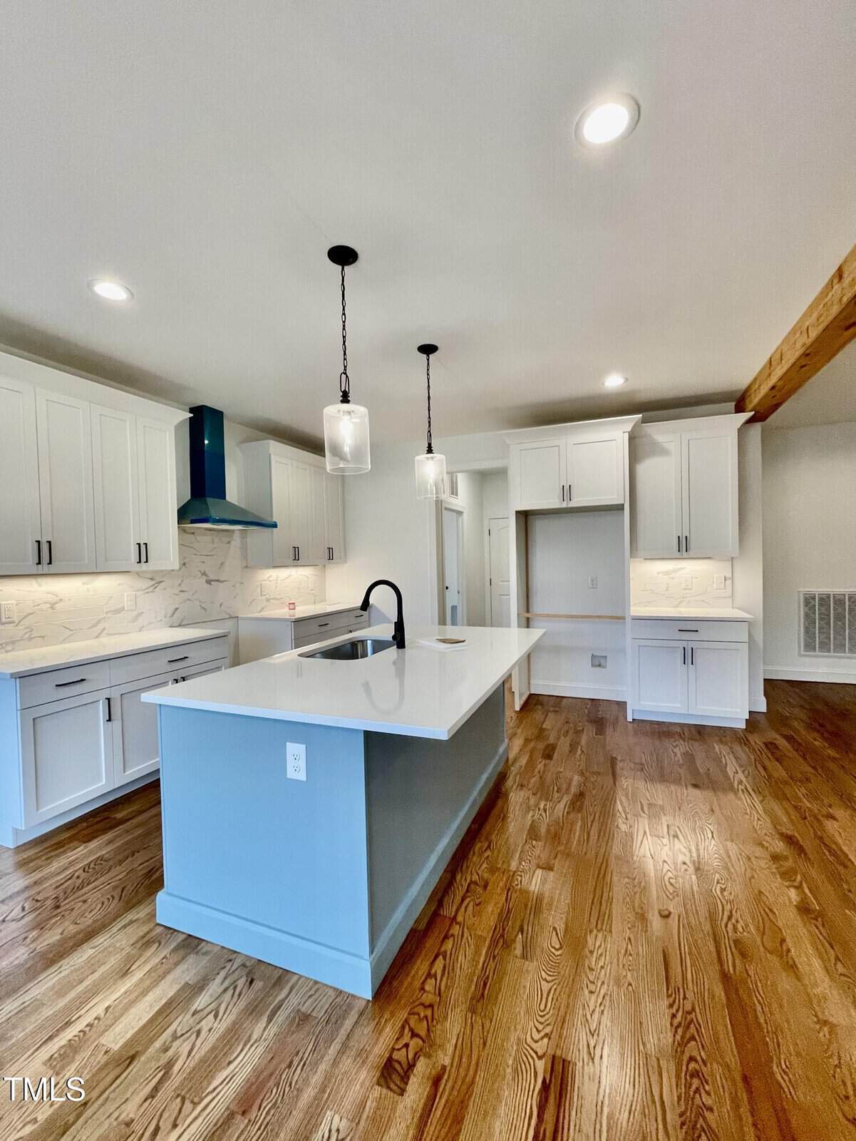 Modern kitchen with white cabinets, a large island with a sink and black faucet, pendant lights, stainless steel range hood, marble backsplash, and wooden floors. The space is bright with recessed ceiling lights.