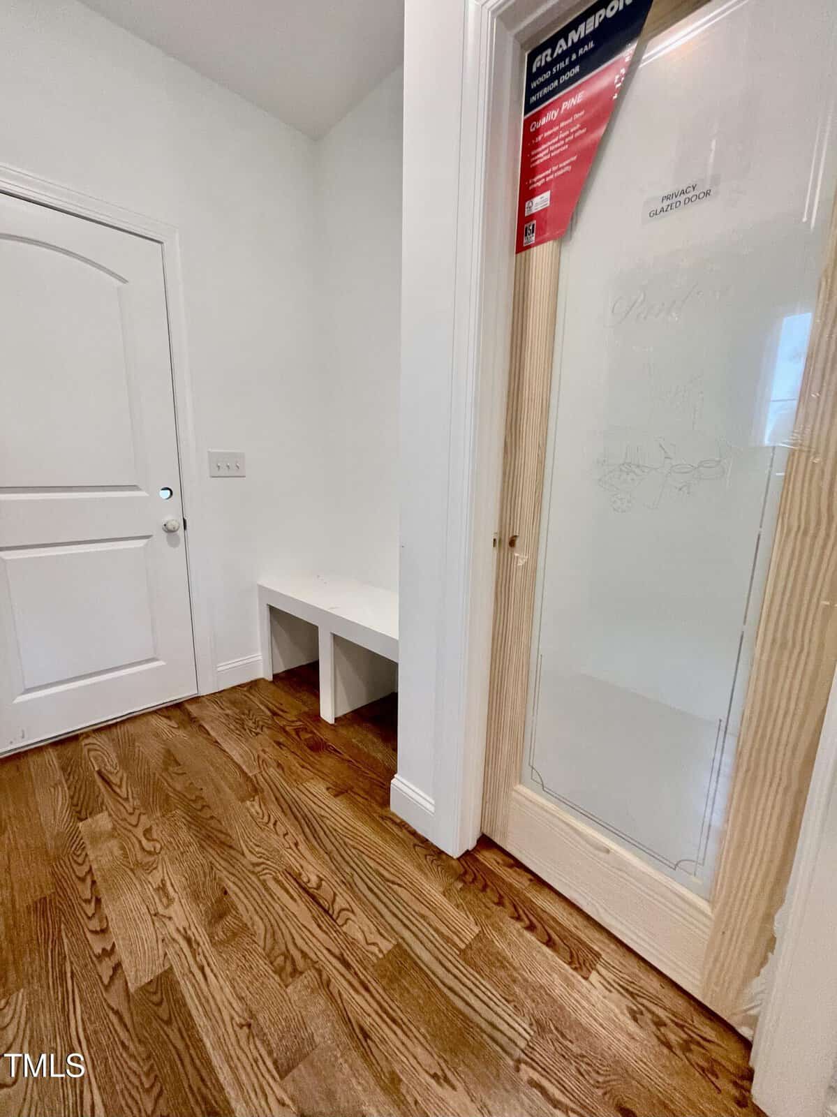 A small entryway with hardwood floors, a white door, a built-in white bench, and a glass pantry door framed with unfinished wood.