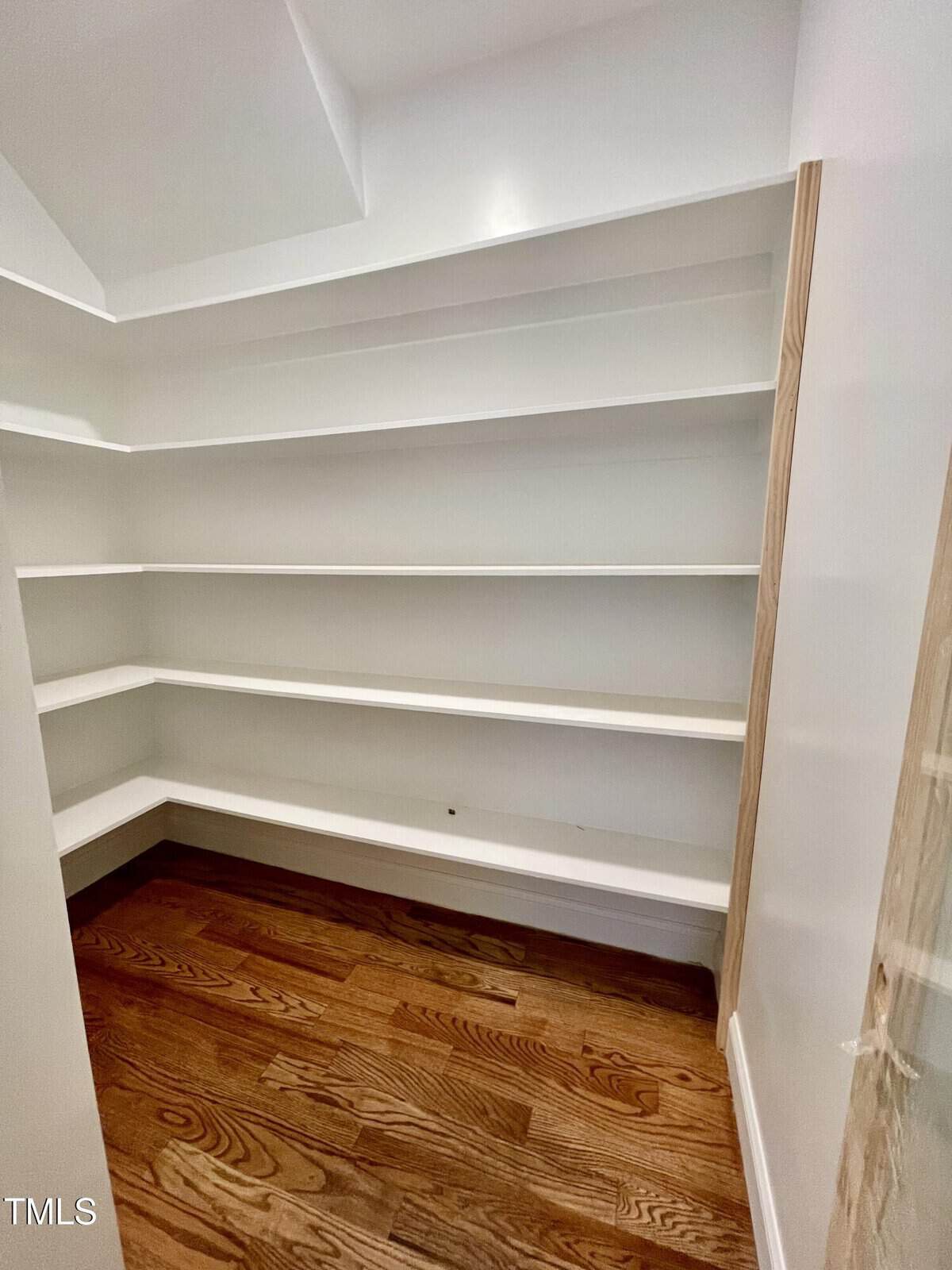 Empty walk-in pantry with white shelves on three walls and hardwood flooring. The shelves are built-in and extend to the corners, providing ample storage space. The room is clean and well-lit.