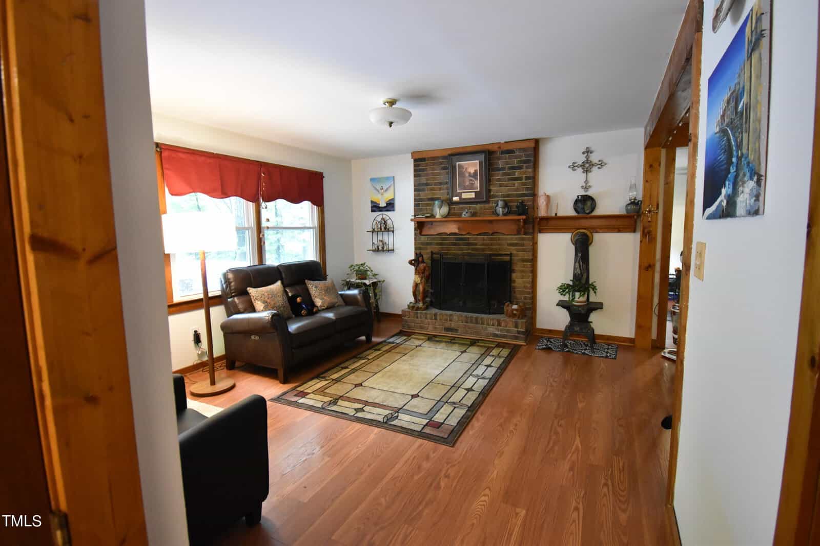 A cozy living room with hardwood floors, a brick fireplace, brown leather seating, a beige patterned area rug, large window with red valance, and wooden trim. Decor includes plants, artwork, and a floor lamp.