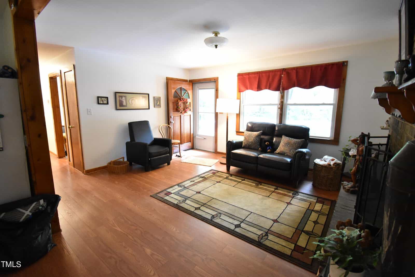 A cozy living room with wood floors, an area rug, a black leather sofa, an armchair, and a basket. Large windows with red valance, a lamp, and a front door let in natural light. A fireplace is visible on the right.