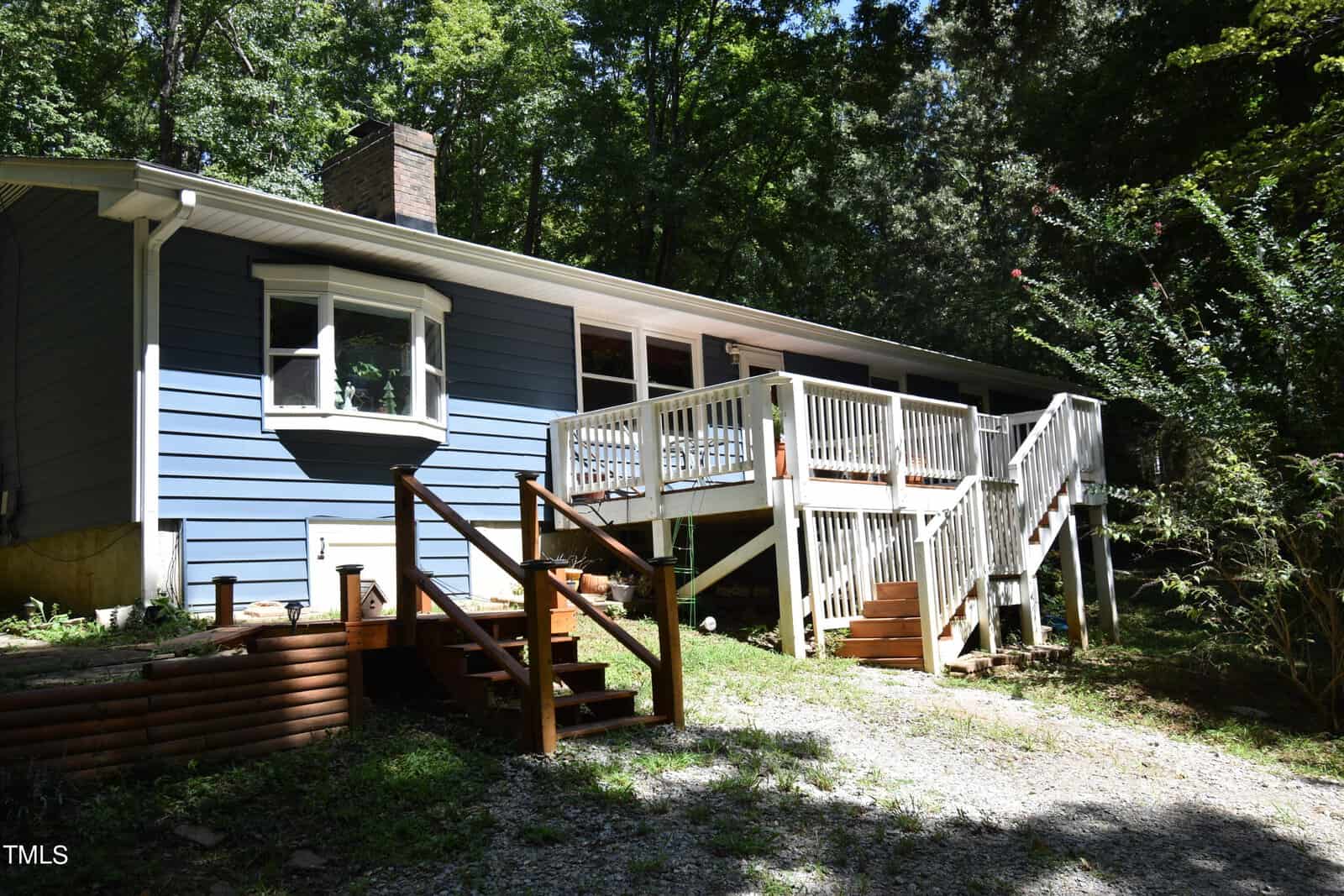 A blue single-story house with white trim, a brick chimney, a bay window, and a large white-railed wooden deck with stairs, surrounded by trees and greenery on a sunny day.