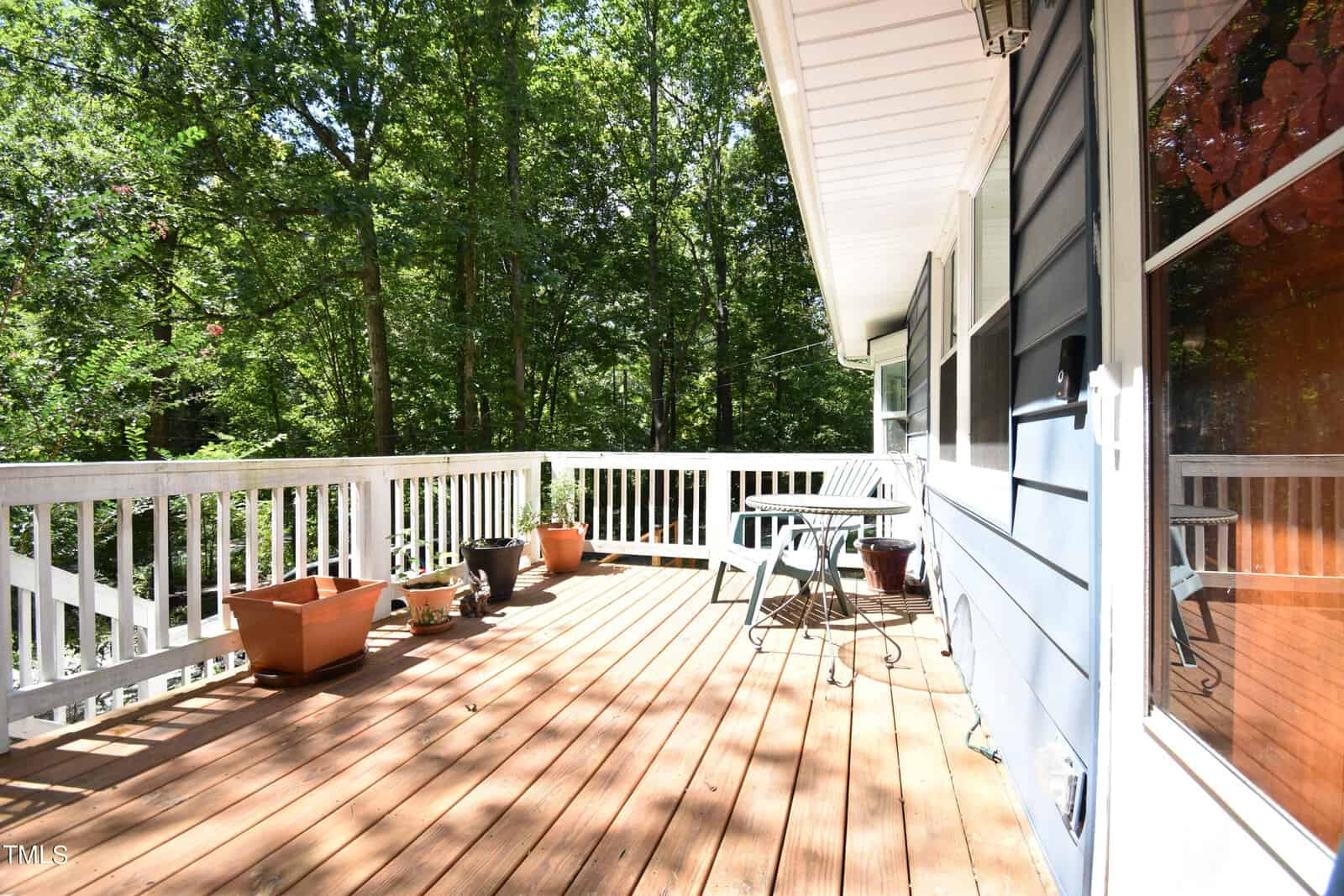 A wooden deck with a white railing features several empty plant pots, a small table, and chairs. The deck is attached to a blue-sided house and overlooks a lush, green forest under bright sunlight.