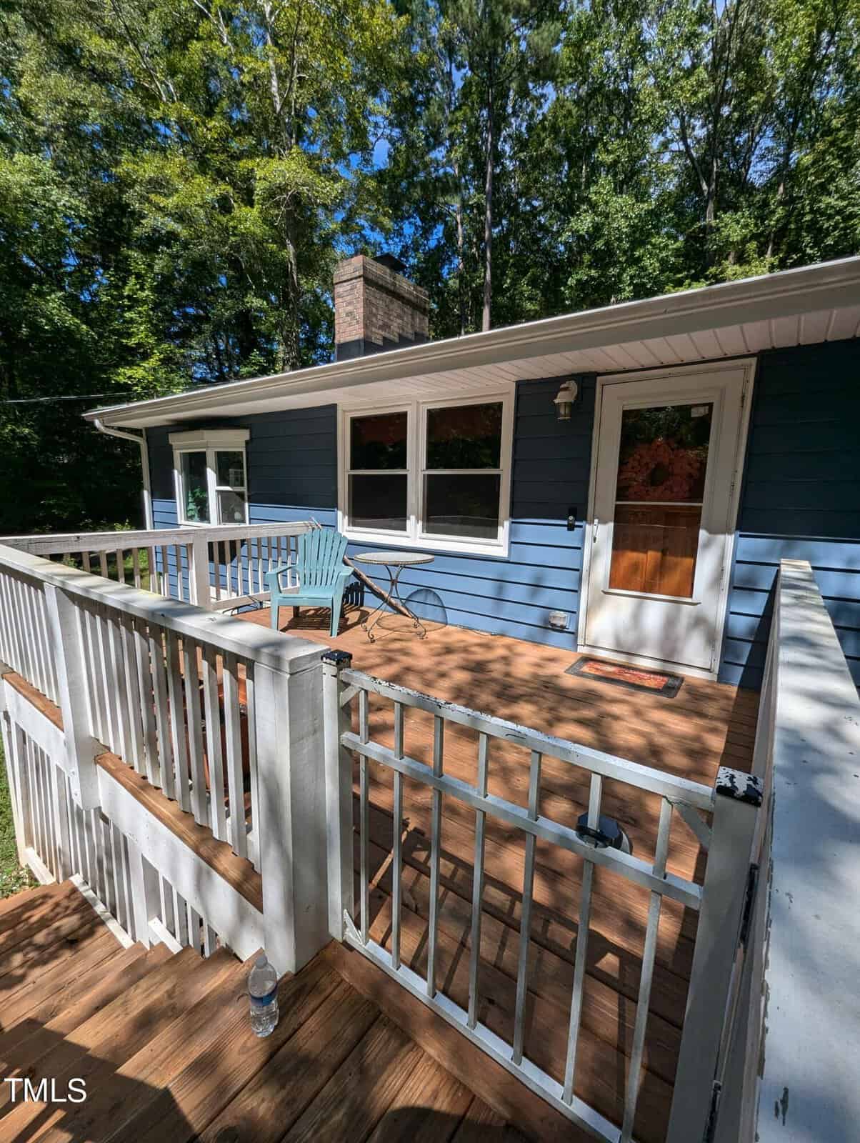 A wooden deck with white railings is attached to a blue house. There is a light blue chair and a small table on the deck. Trees and greenery surround the house in the background.