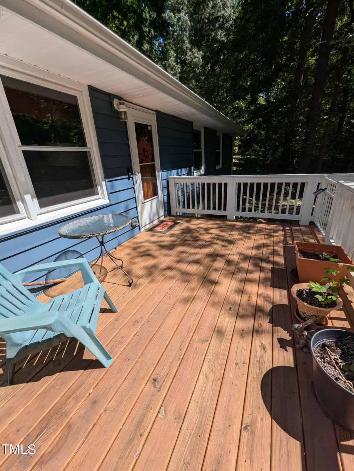 A wooden deck with a glass table, blue chair, and potted plants, attached to a blue house with white trim, overlooks a wooded area. Sunlight casts shadows on the deck.