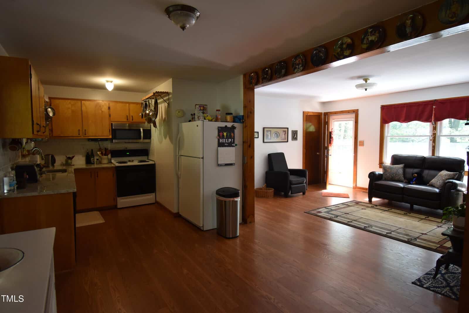 Open kitchen and living area with wooden floors, white appliances, brown cabinets, a small sitting area with a black couch and armchair, a large window, and decorative plates on a wooden beam.