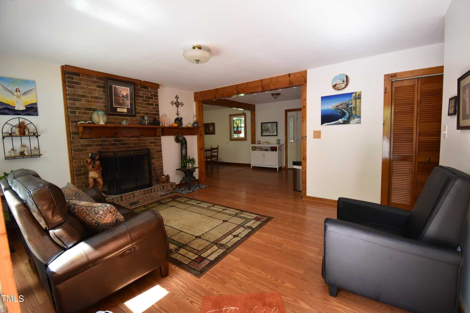 A cozy living room with wood floors, a brown leather sofa, a black armchair, a brick fireplace with a wooden mantle, a rug, and wall art. An open doorway leads to another room with more art and furniture.