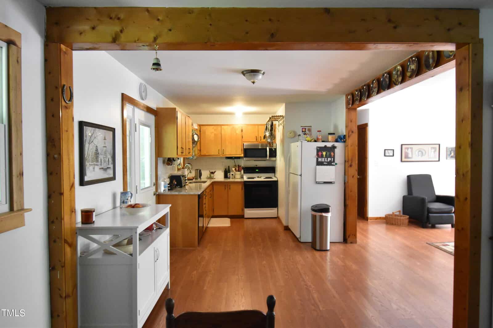 A cozy kitchen with wooden cabinets, white appliances, and hardwood floors, viewed from a dining area. Light streams in through windows, and decorations line the walls and shelves.