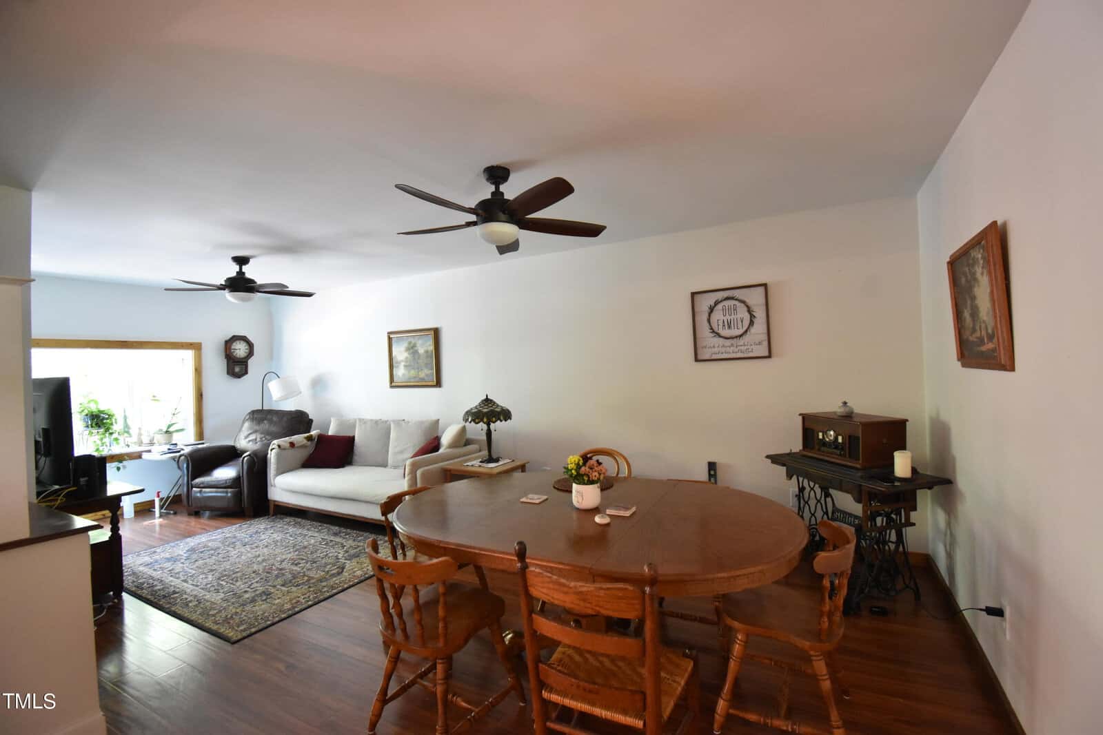 A living and dining area with wooden floors, a round wooden dining table with chairs, a sofa, armchair, side tables, artwork on white walls, ceiling fans, and a window letting in natural light.