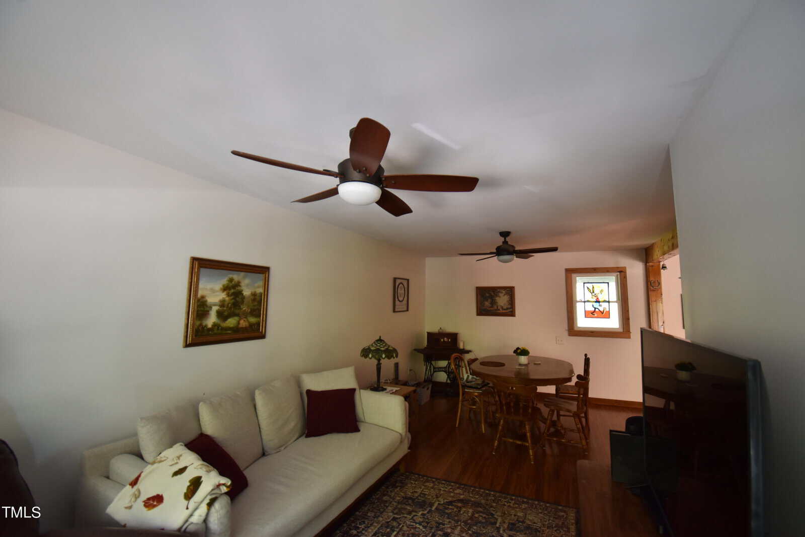 A living room with a beige sofa, paintings on the wall, a round dining table, ceiling fans, a stained glass window, and wooden floors. A TV is angled in the corner on the right.