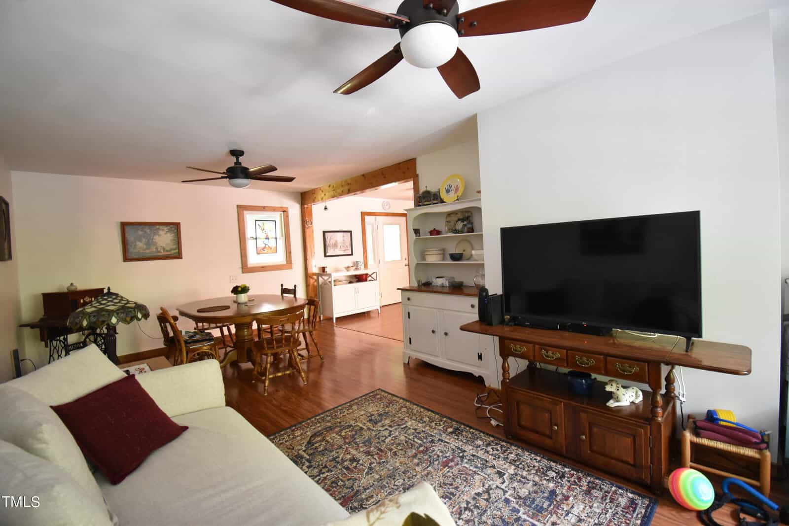 A cozy living room with a beige sofa, patterned rug, and large TV on a wooden console. The room has wood floors, ceiling fans, a dining area with table and chairs, and an open view into a bright kitchen.