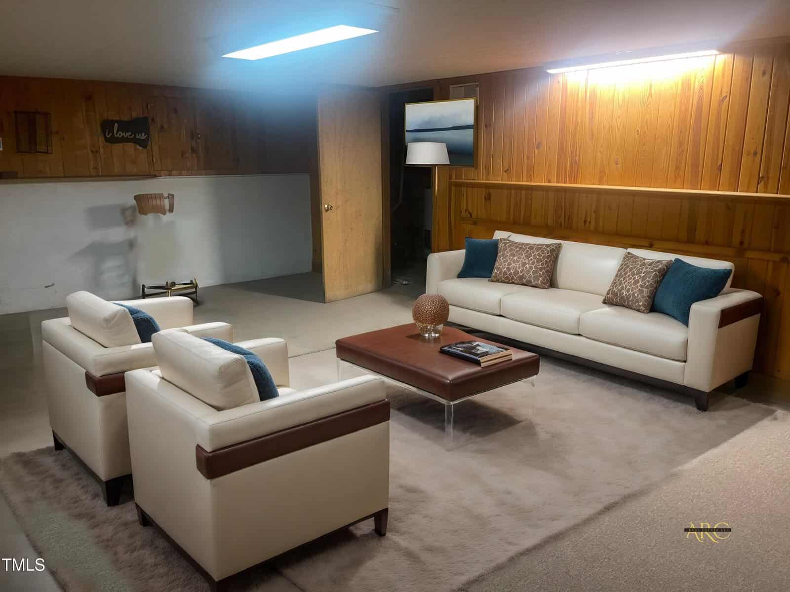A cozy basement living room with wood-paneled walls, featuring a white sofa and two matching armchairs with blue accents. A brown coffee table is centered with decor. A standing lamp and artwork on the wall add warmth to the space.