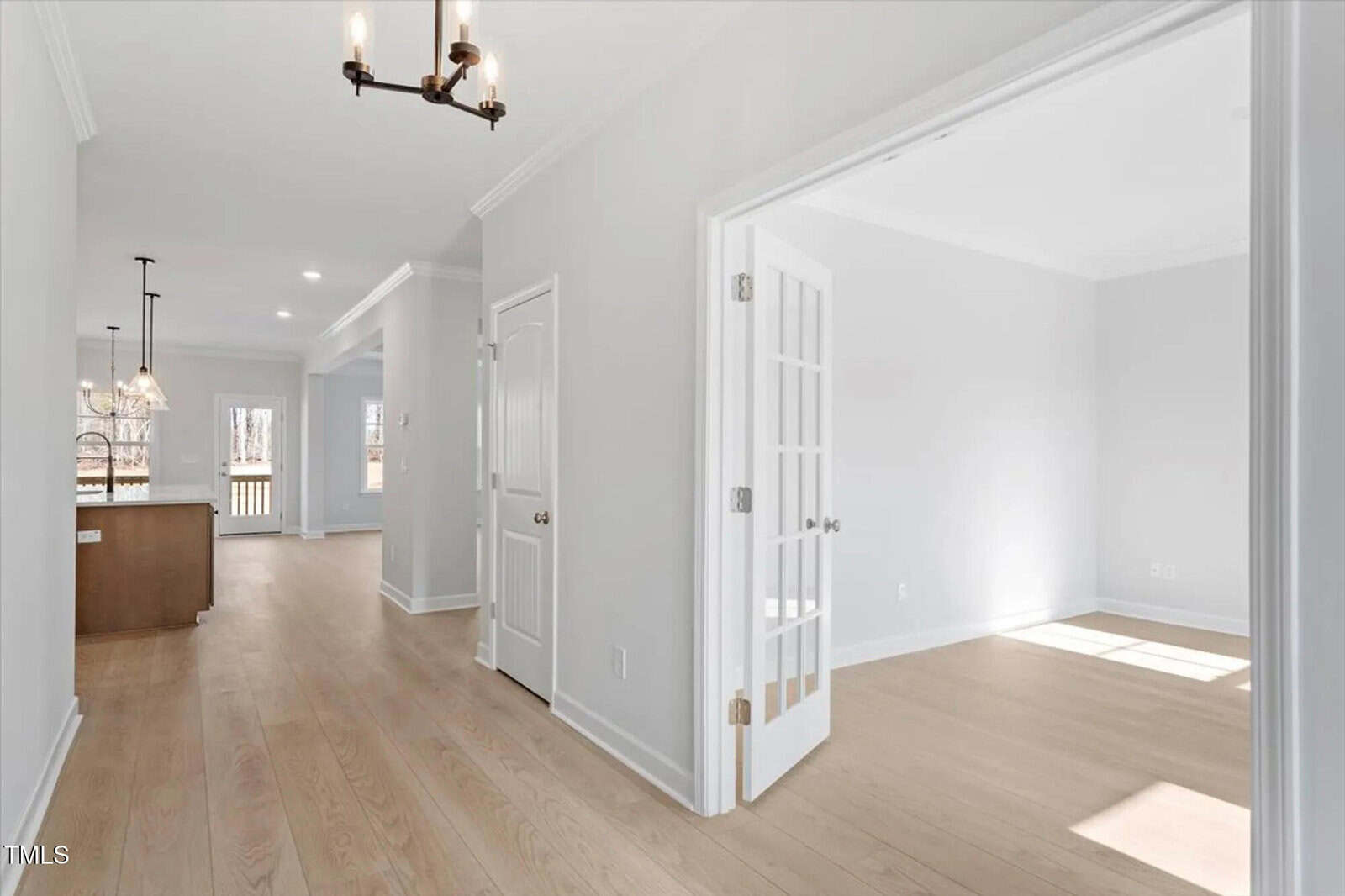 Bright hallway in a modern home with light wood floors, white walls, and a chandelier. French doors open to a sunlit room on the right; kitchen with pendant lights and deck visible in the background.