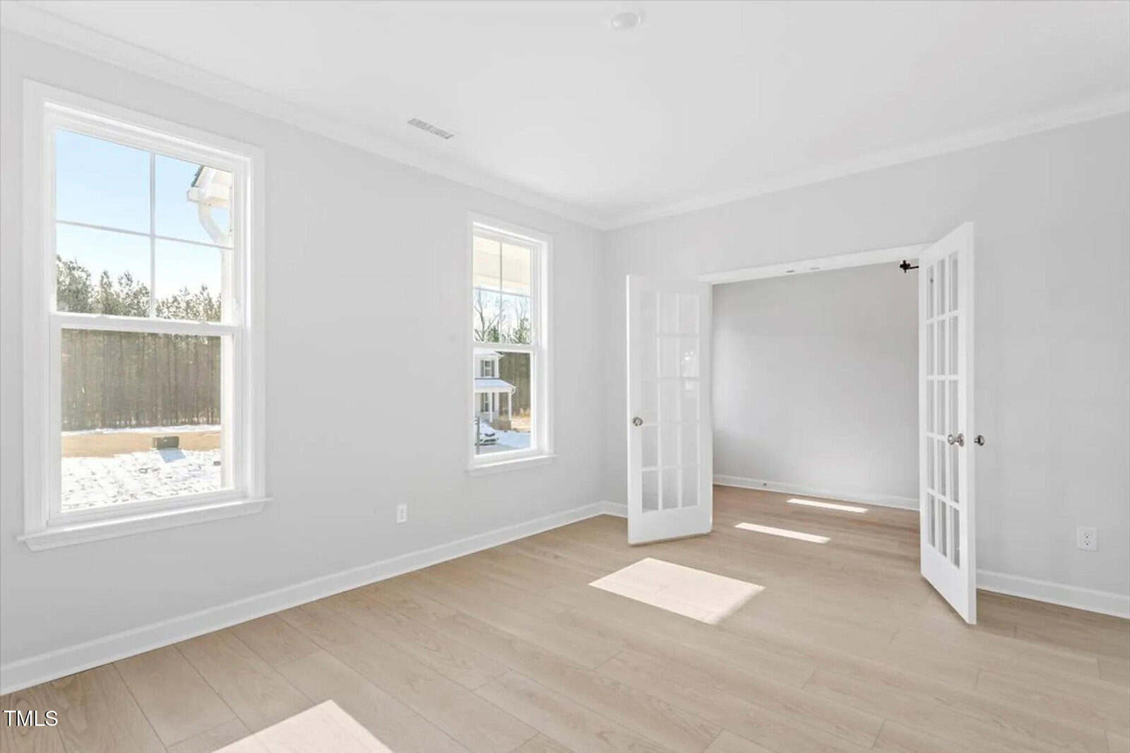 Bright, empty room with light wood flooring, two large windows, and white walls. Sunlight streams in. French doors open into another room. A fenced yard with snow is visible through the windows.