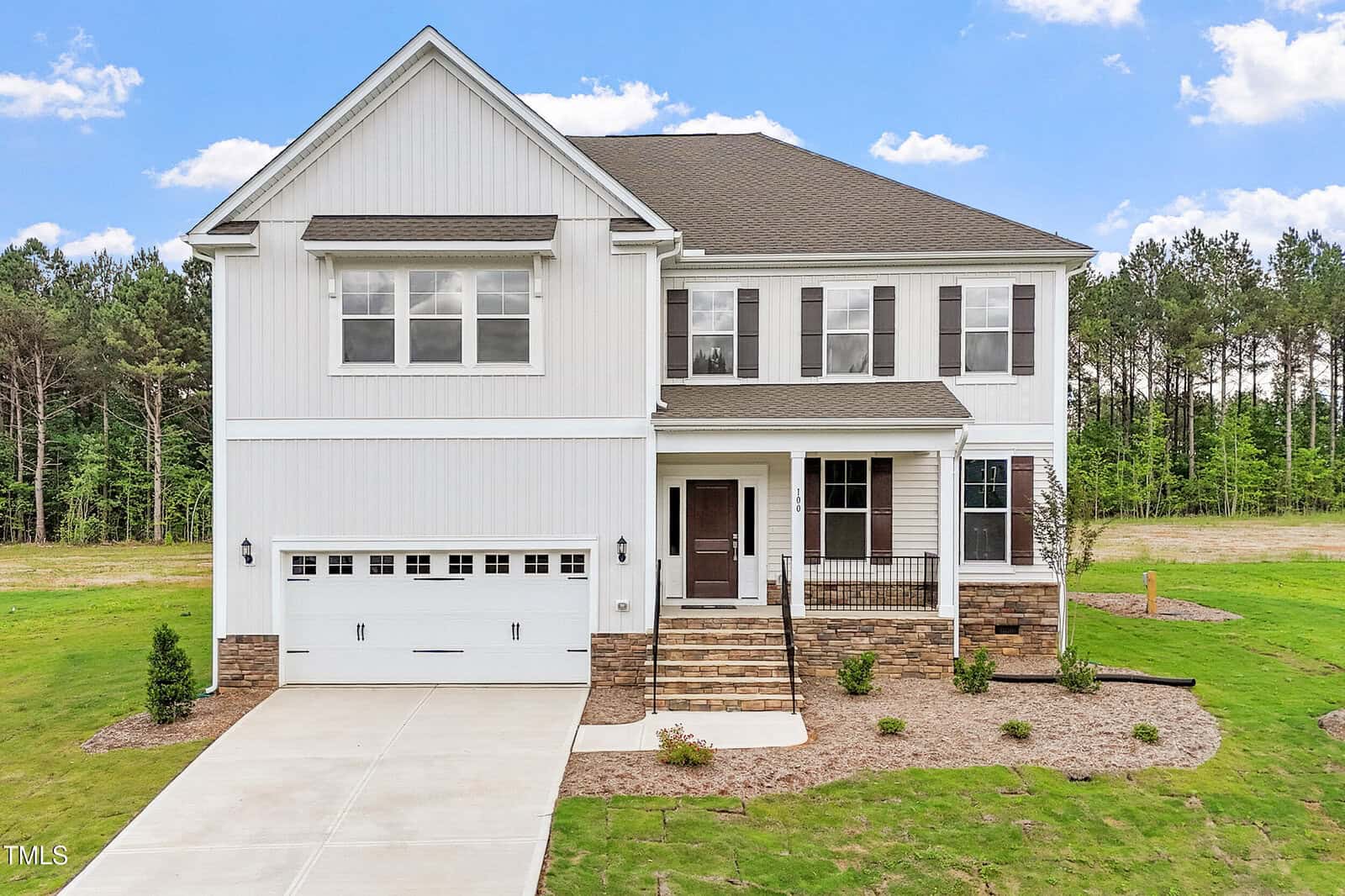 Located at 100 Bold Drive in Youngsville, this two-story white house features a dark roof, spacious two-car garage, inviting front porch, and steps leading to the entrance. The yard is grassy with tidy mulch beds and small plants, while mature trees and a blue sky with clouds create a picturesque backdrop.