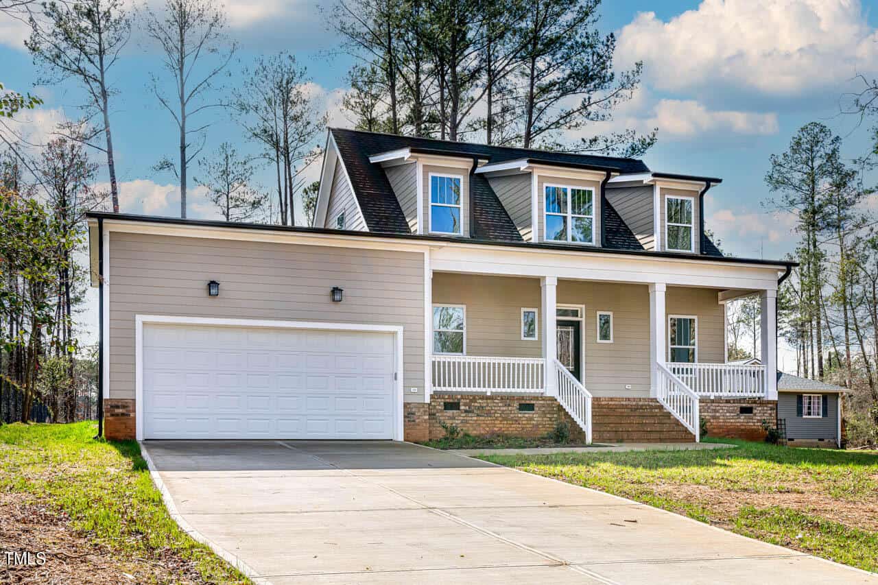 A two-story house with beige siding, white trim, three dormer windows, a covered front porch with white railings, a double garage, and a concrete driveway, surrounded by trees.
