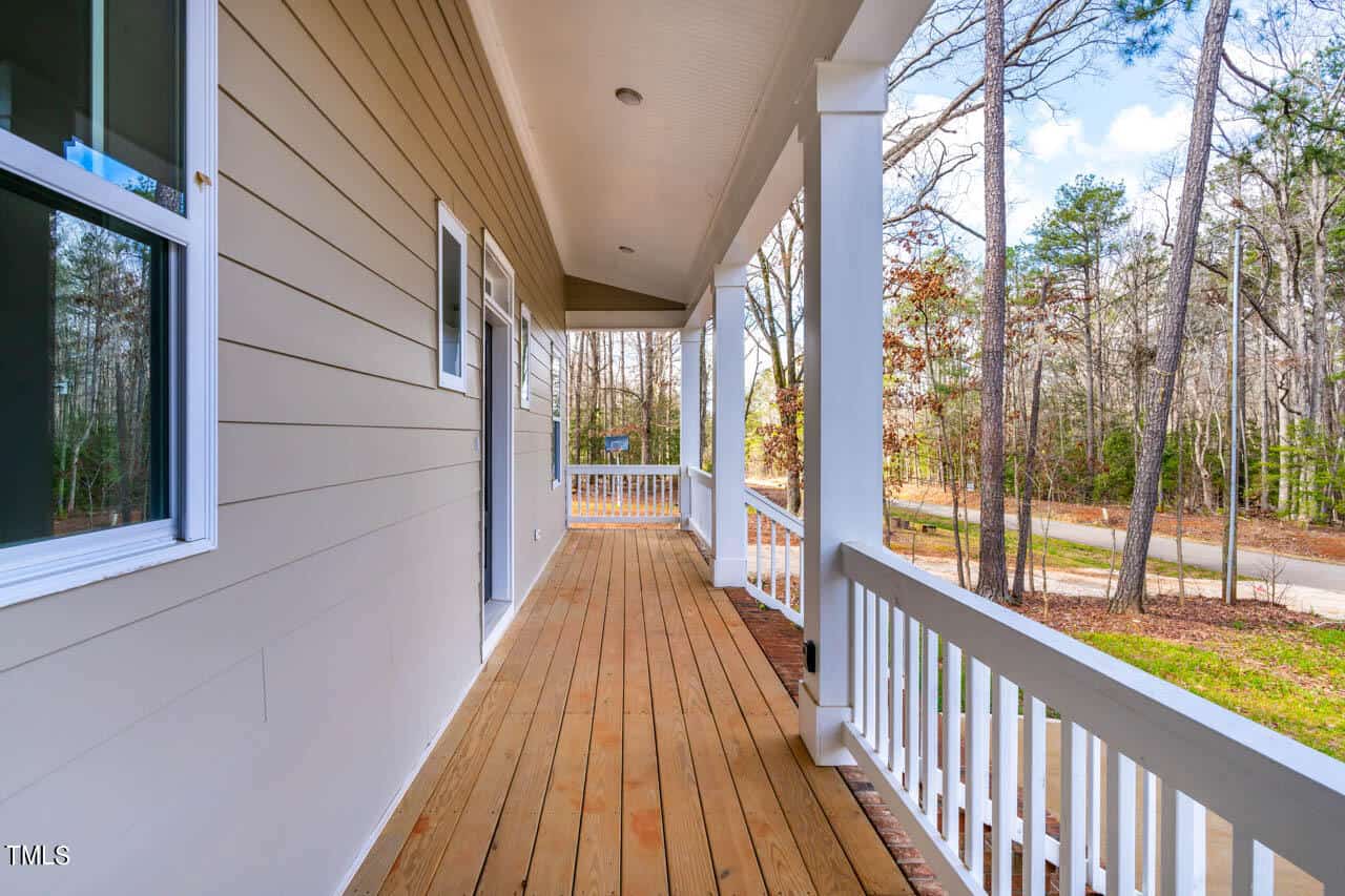 A wooden porch with white railings runs along the side of a house with beige siding. Trees and a street are visible in the background. The porch is covered by a roof with recessed lighting.