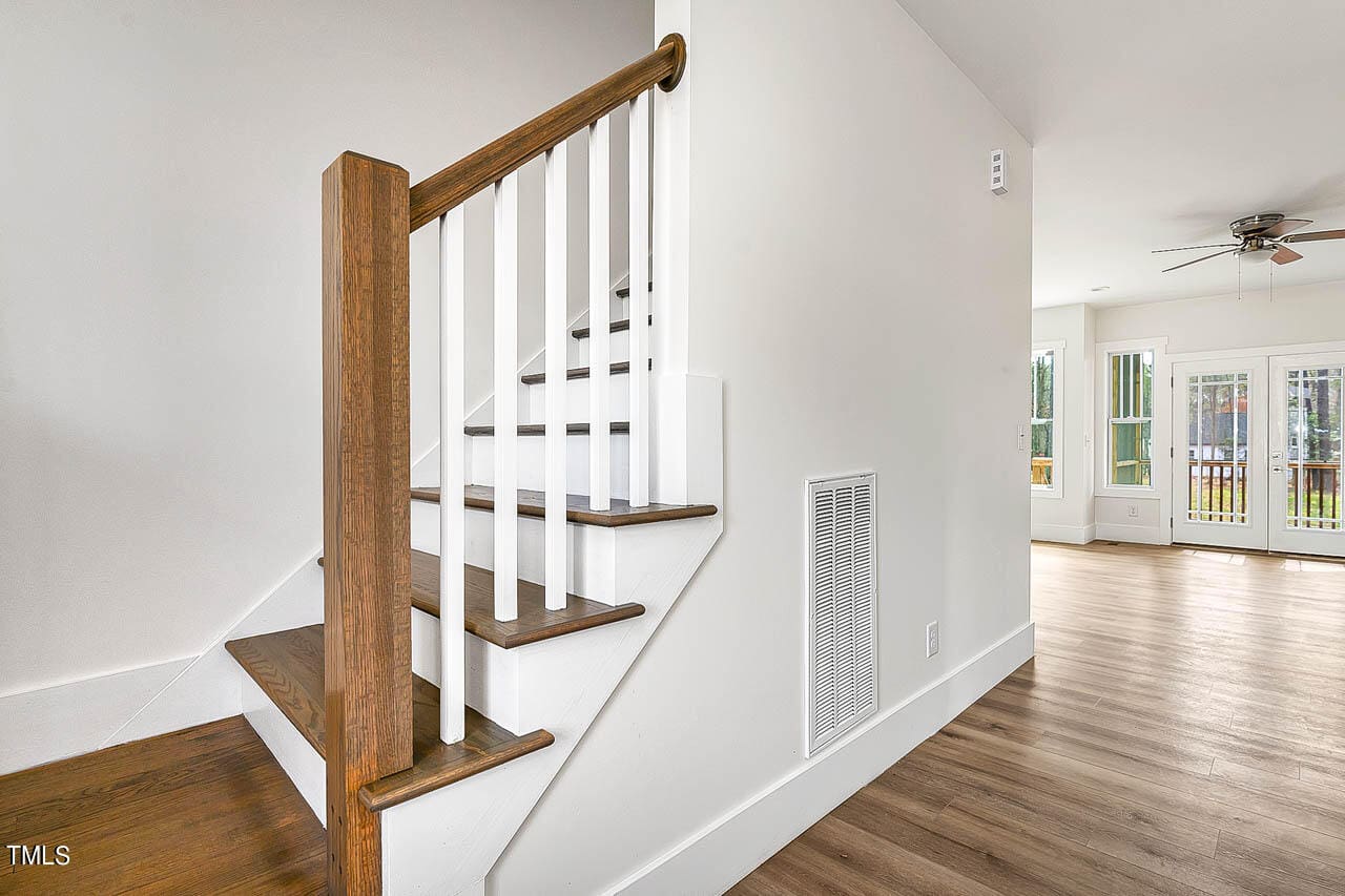 A wooden staircase with white railings leads upstairs in a modern home with white walls and light wood floors. A living area with ceiling fan, windows, and glass doors is visible in the background.