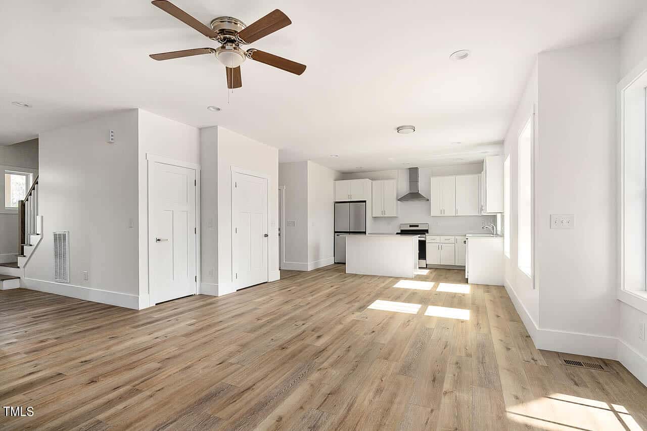 Bright, modern open-concept living area with wood floors, white walls, ceiling fan, kitchen with white cabinets, stainless steel appliances, island, and multiple doors; staircase visible on the left.