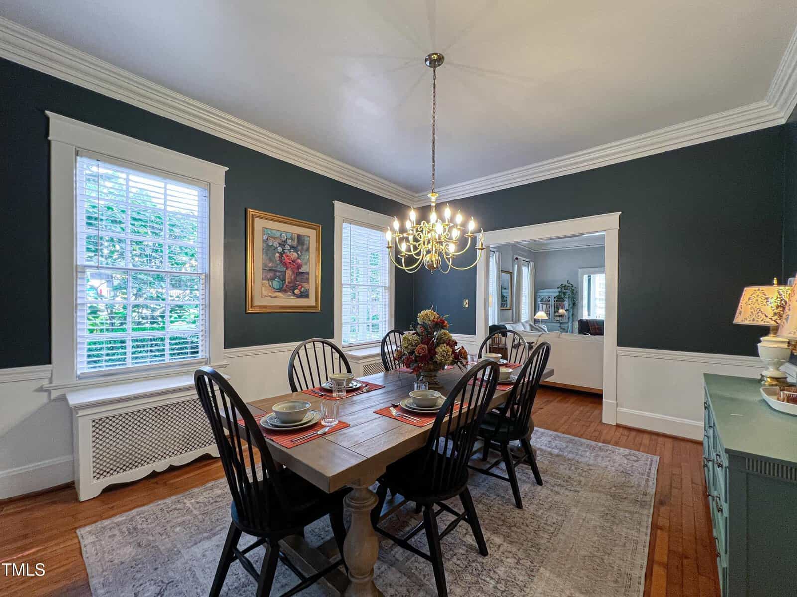 A dining room with dark green walls and white wainscoting features a wooden dining table set for six with black chairs. A large chandelier hangs above the table. A painting is on the wall, and through an opening, more of the house is visible. Natural light enters through two tall windows.