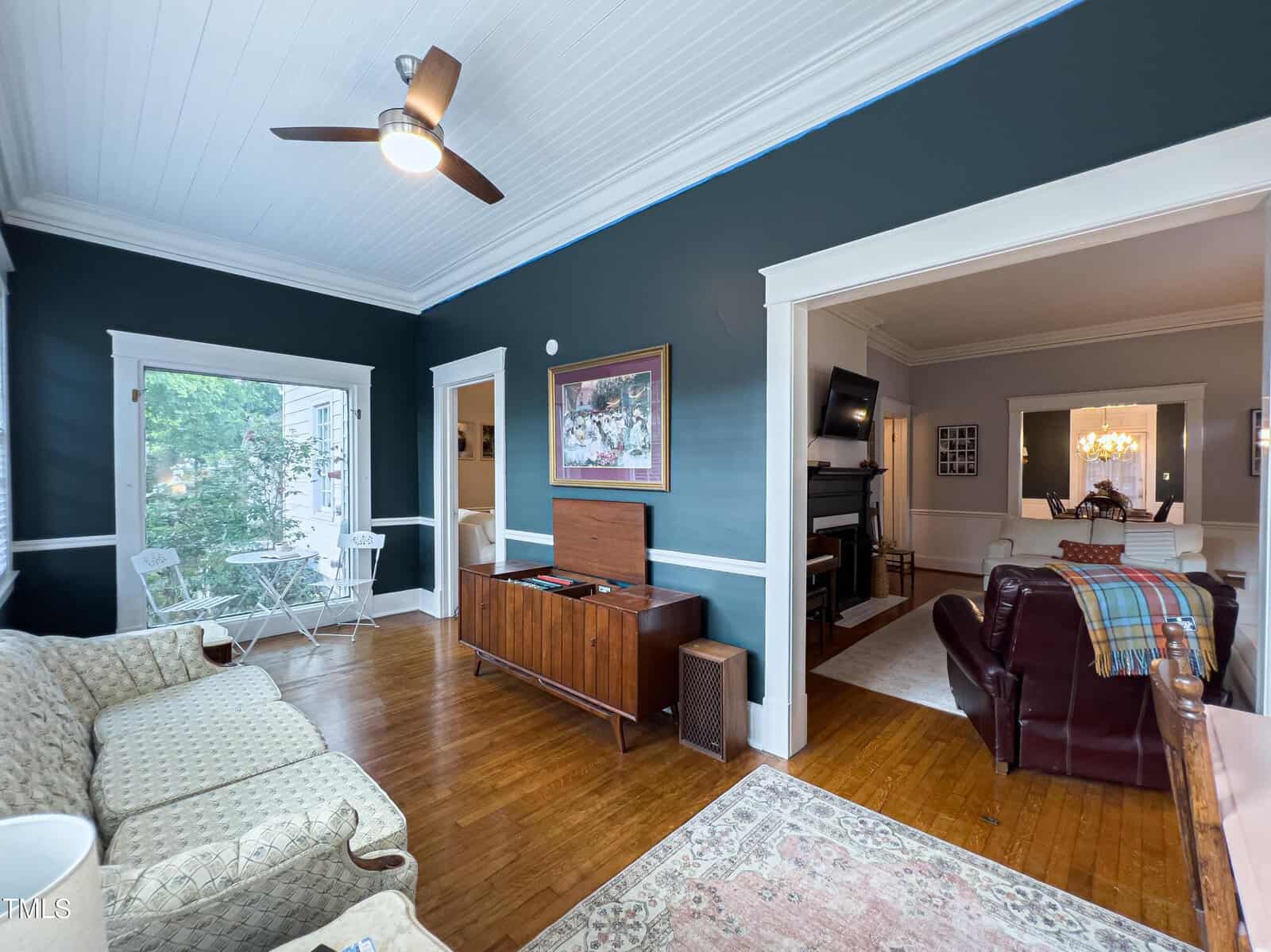 A cozy living room features dark green walls, a wooden floor, and white trim. It includes a ceiling fan, a vintage-style sofa, an area rug, and a wooden stereo cabinet. An adjoining room with a chandelier is visible through a wide doorway. Large windows bring in natural light.