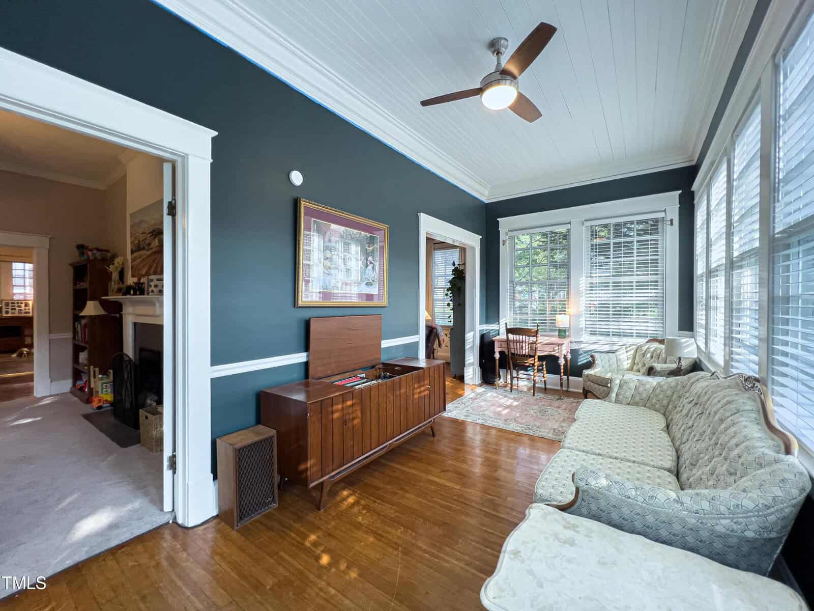 A sunlit living room with dark teal walls, white trim, and wooden flooring. The room features a ceiling fan, a mixed-style seating arrangement, and vintage furniture including a sofa, armchair, and a wooden cabinet with a record player. Large windows allow natural light to flood the space.