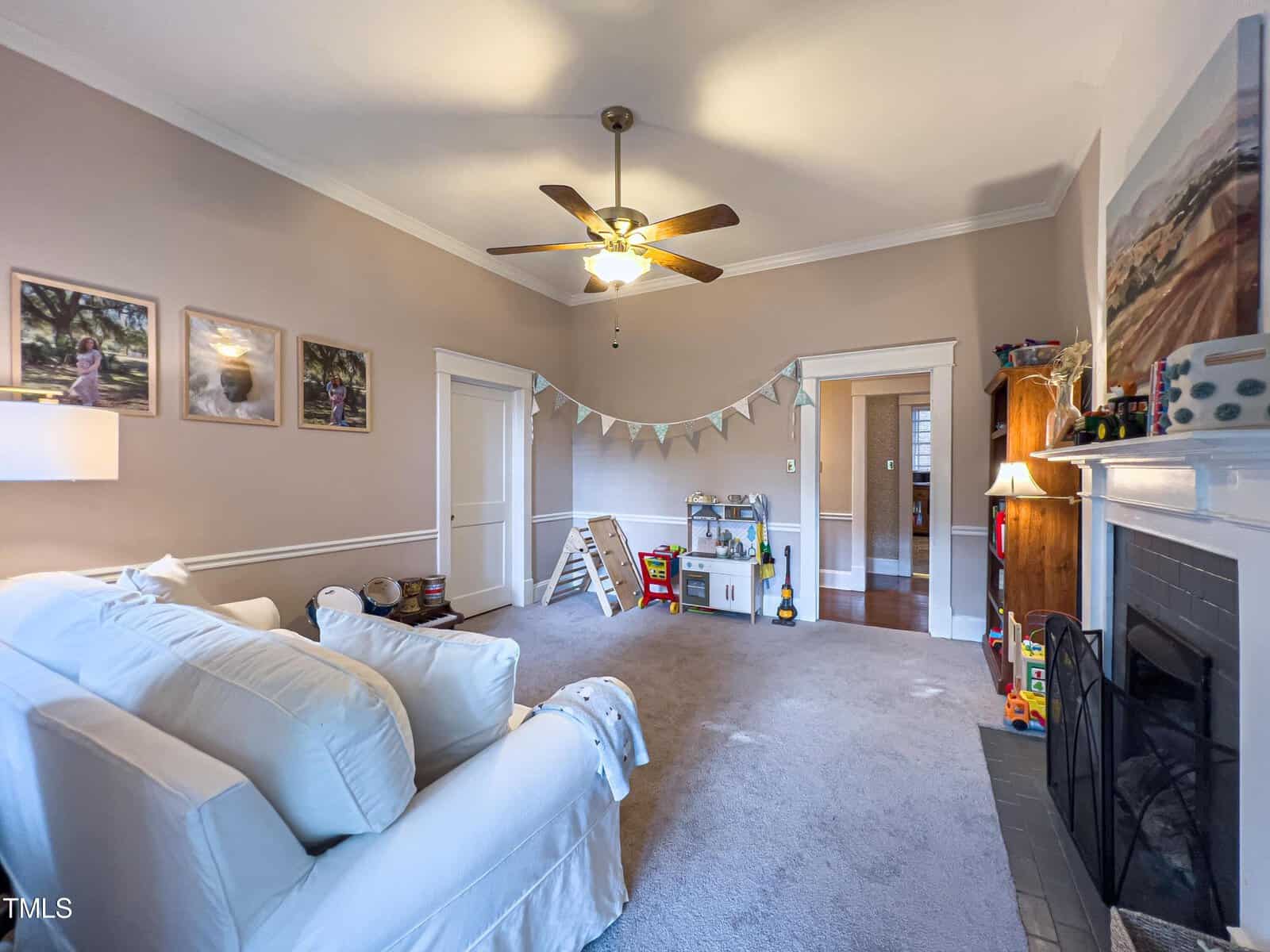 A cozy living room with a white couch, ceiling fan, and beige walls adorned with framed pictures. A small play area with toys and a white bookshelf is set up near the fireplace. A decorated banner hangs above the play area, and a door leads to another room.