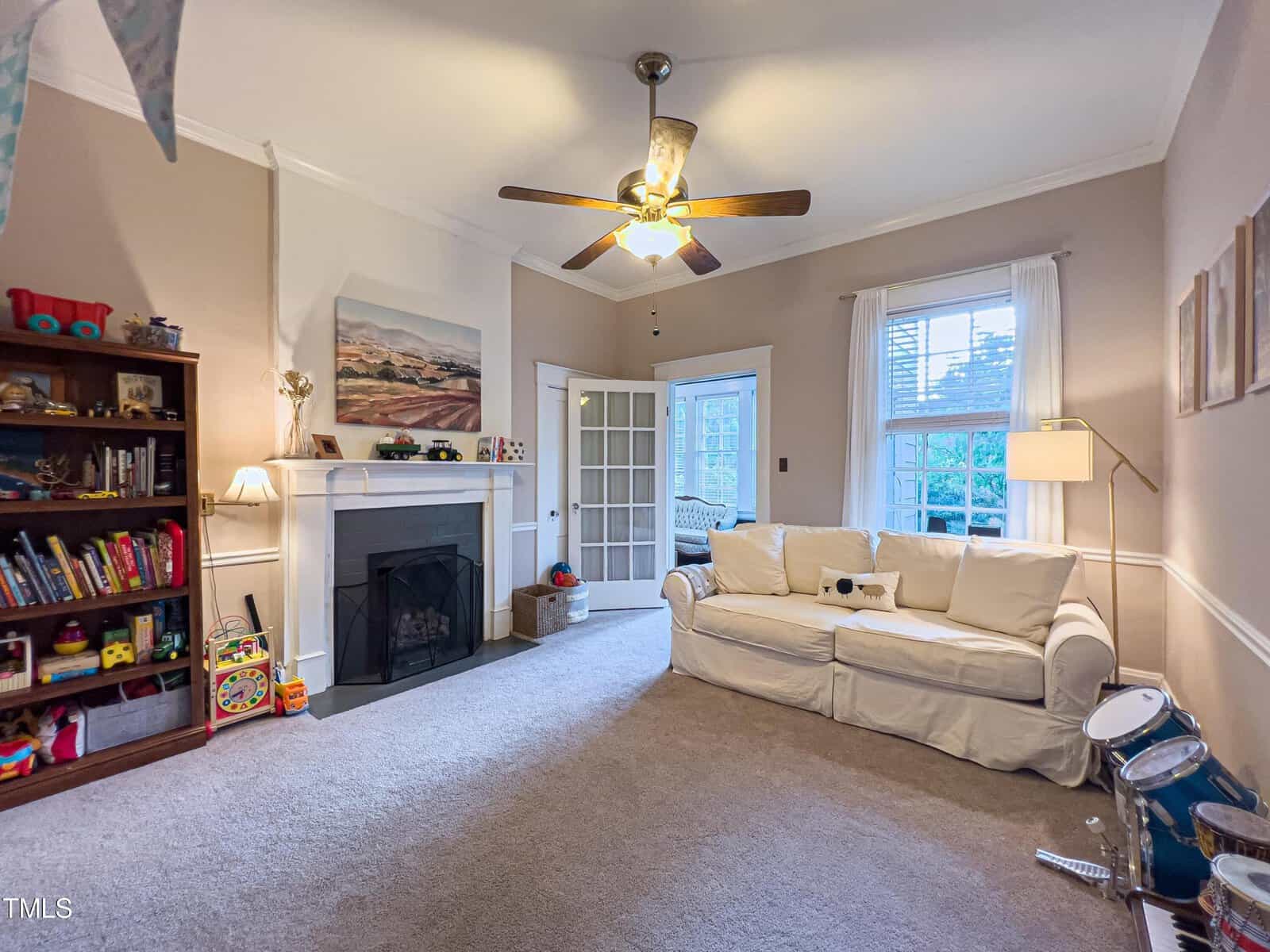 A cozy living room with beige walls and a ceiling fan. A white sofa sits next to a floor lamp and small drum set. A bookshelf filled with toys is located by the fireplace. A doorway leads to another room with a white sofa and windows. A framed picture hangs above the fireplace.