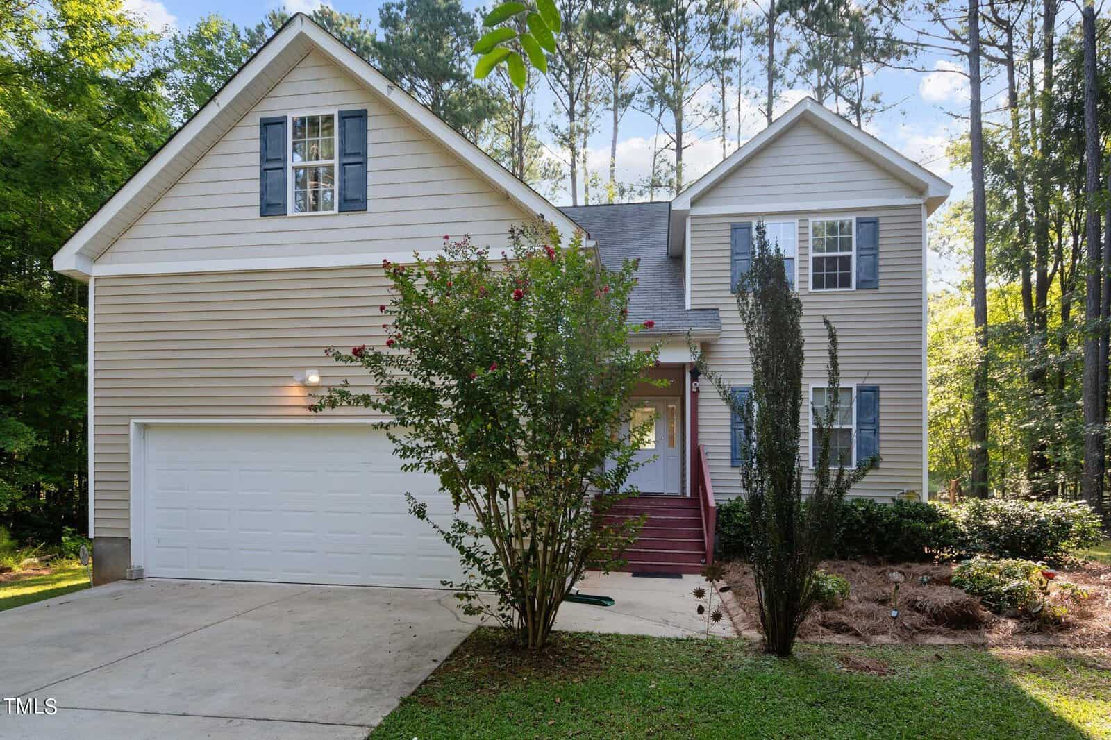 A beige two-story suburban house with blue shutters, a double garage, red steps leading to the front door, and landscaping with shrubs and trees, surrounded by tall pine trees.