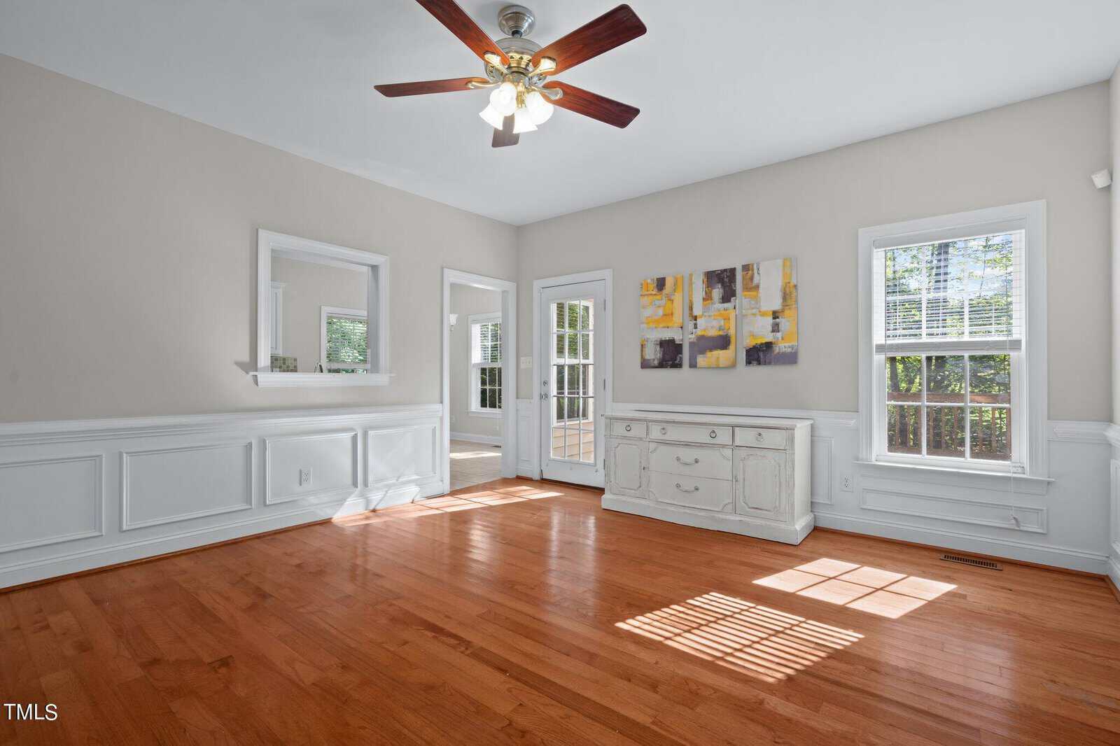 Bright room with hardwood floors, white wainscoting, ceiling fan with lights, two large windows, French doors, wall mirrors, a sideboard, and abstract artwork on the wall. Sunlight streams through the windows.