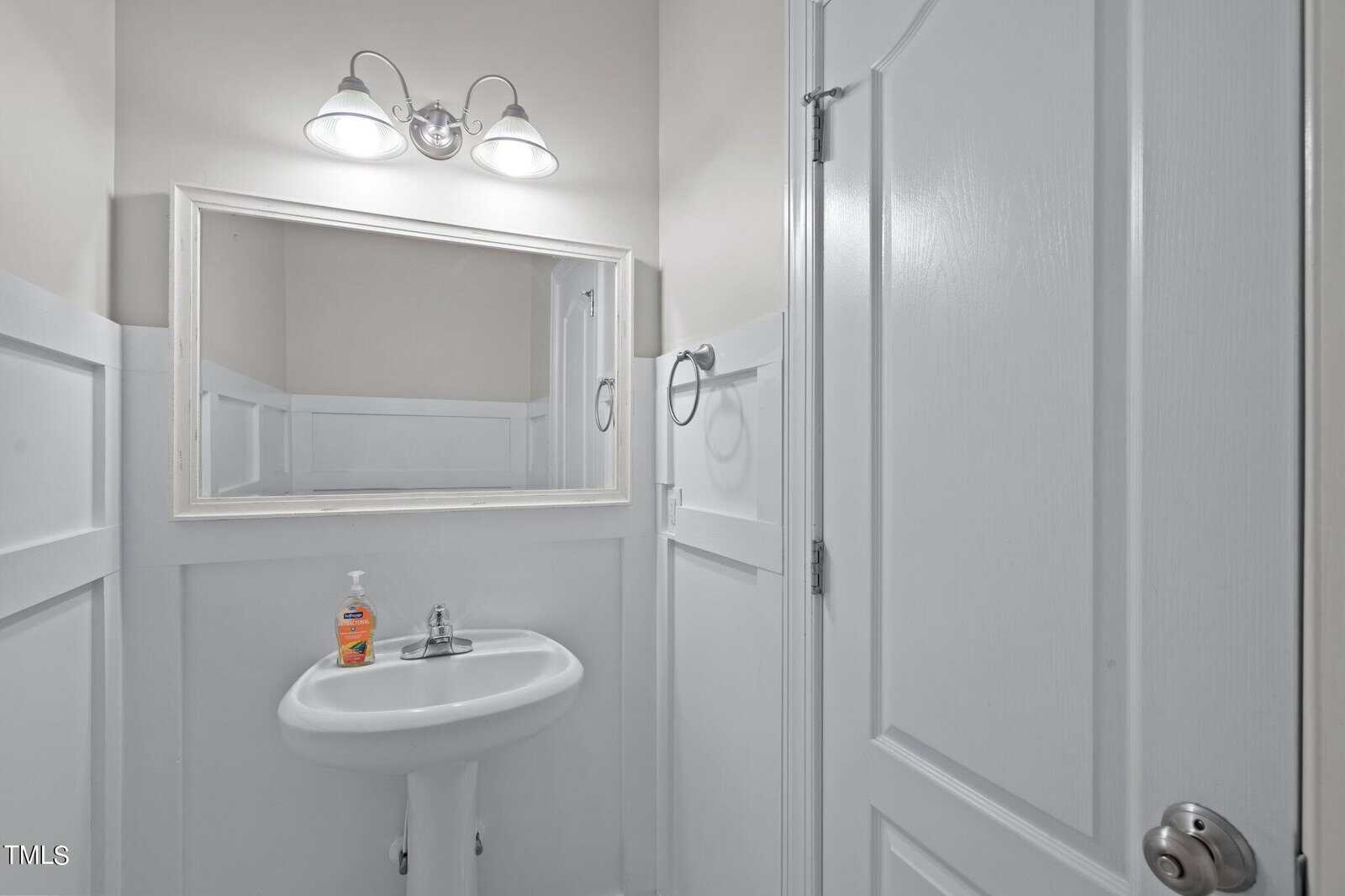 A small, white bathroom with a pedestal sink, a rectangular mirror, and wall-mounted lights. A bottle of hand soap sits on the sink. The walls have white panel wainscoting and there is a towel ring on the right.