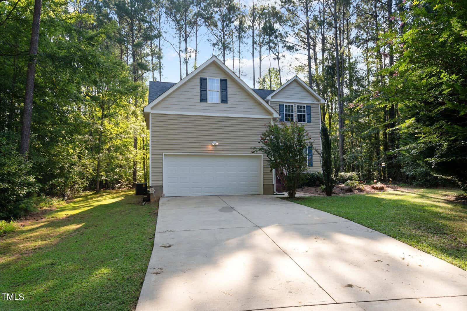A two-story beige house with a white garage door sits at the end of a long driveway, surrounded by tall trees and greenery on a sunny day.