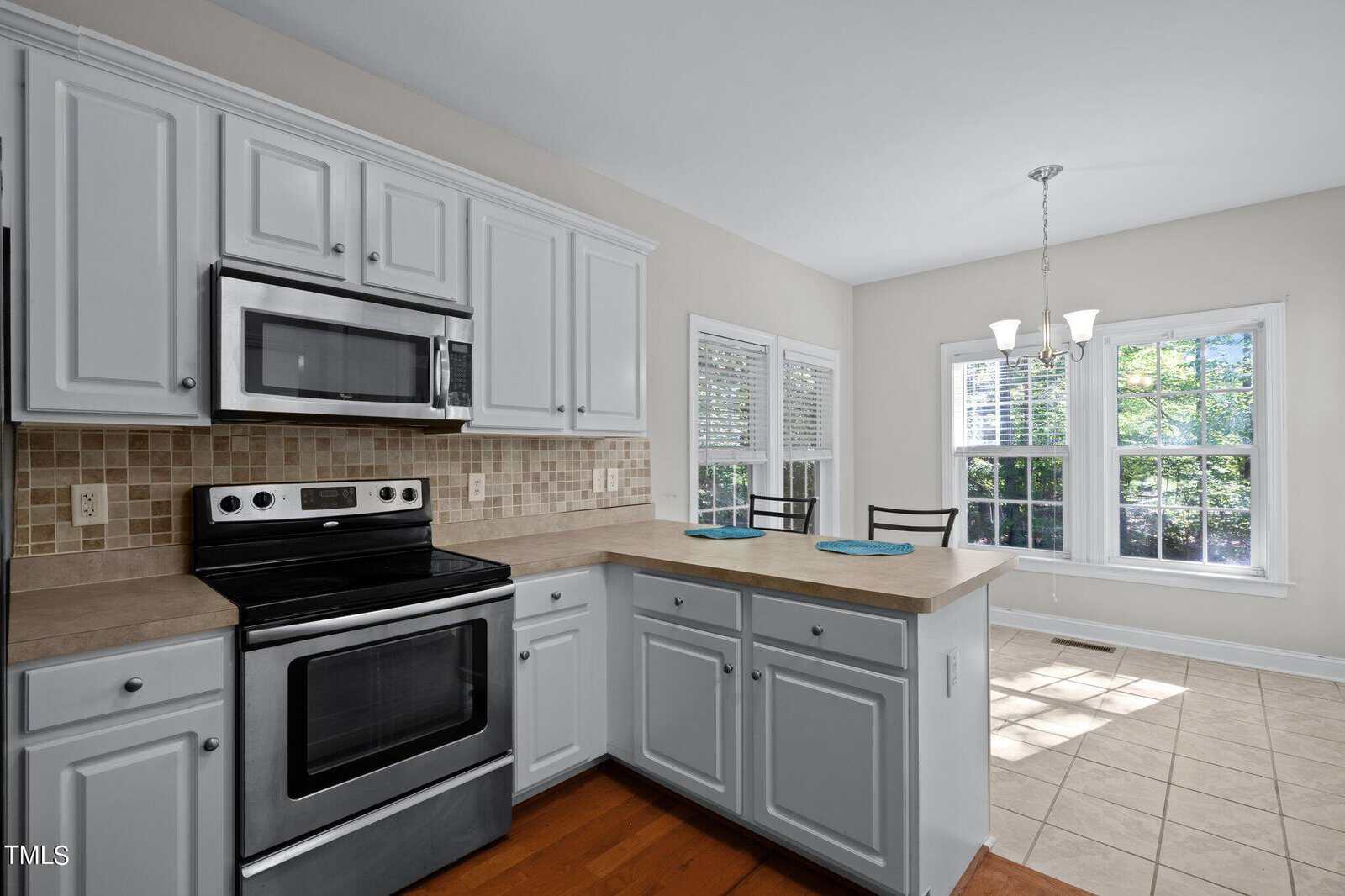 A bright kitchen with white cabinets, stainless steel oven and microwave, tile backsplash, and a breakfast bar. Large windows and a light fixture illuminate the adjacent dining area with tiled floors.