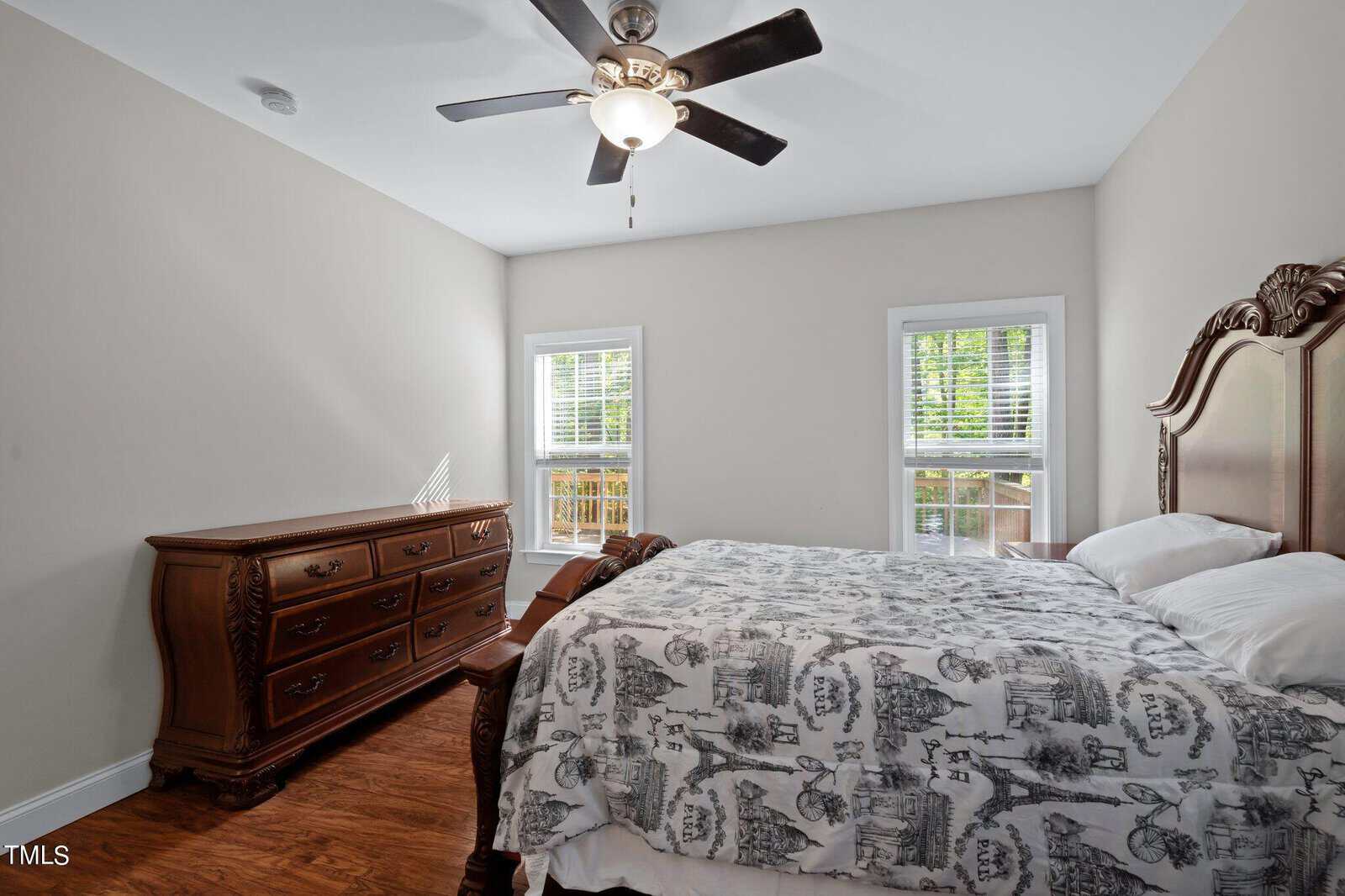 A bedroom with a ceiling fan, two windows with blinds, a wooden dresser, and a matching wooden bed with white and gray patterned bedding. The walls are light beige and the floor is hardwood.