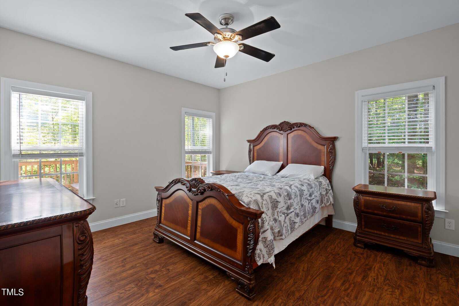 A bright bedroom with wood floors, a ceiling fan, two large windows, an ornate wooden bed with a patterned quilt, a matching nightstand, and a dresser. Green trees are visible outside the windows.