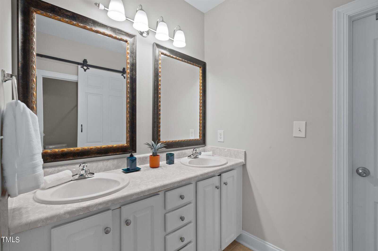 A bathroom with double sinks, two mirrors, white cabinets, towel, soap dispensers, a small plant, and a sliding barn door reflected in the mirrors beneath three wall lights.