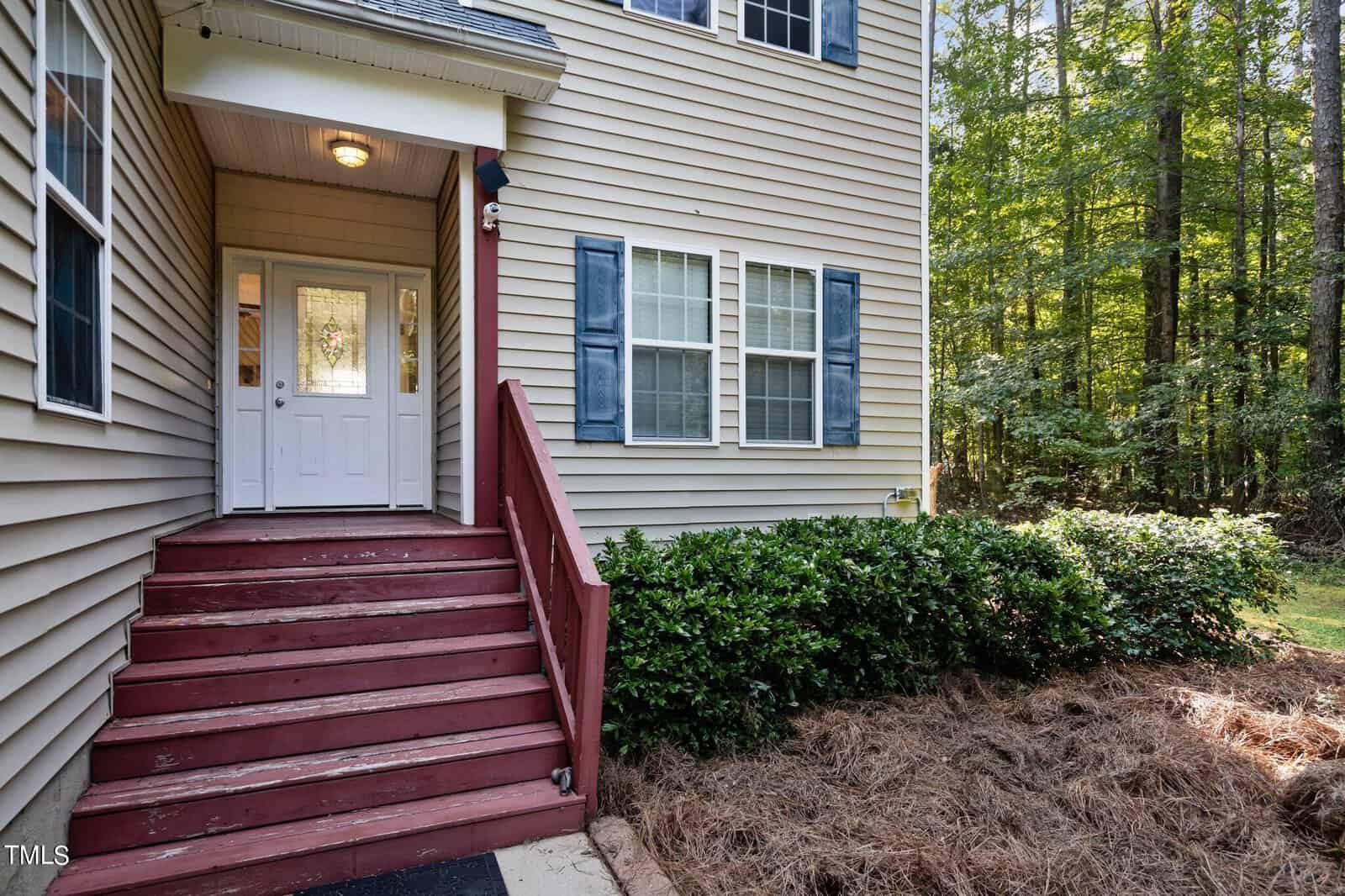 Front entryway of a beige house with red wooden steps, white double doors, and blue shutters beside windows. Shrubs and pine straw line the walkway; tall trees are visible in the background.