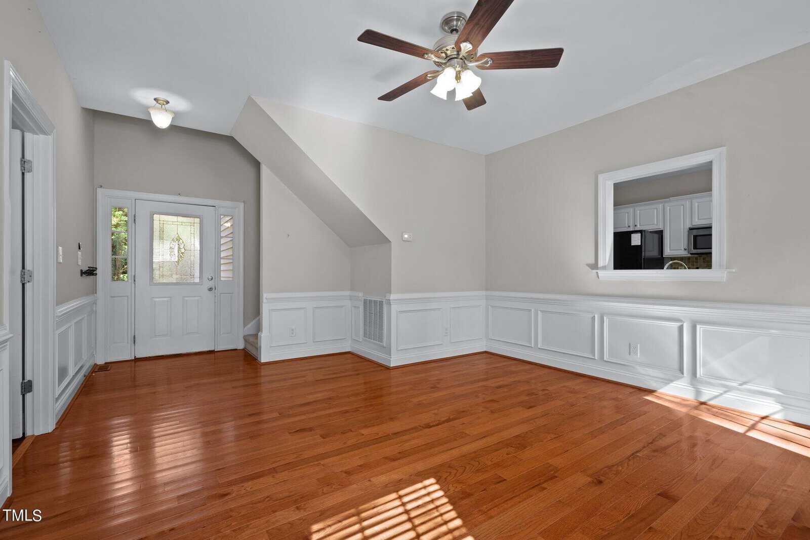 Bright, empty room with hardwood floors, white wainscoting, beige walls, ceiling fan with lights, and a window opening to a kitchen. Front door and small window are visible in the background.