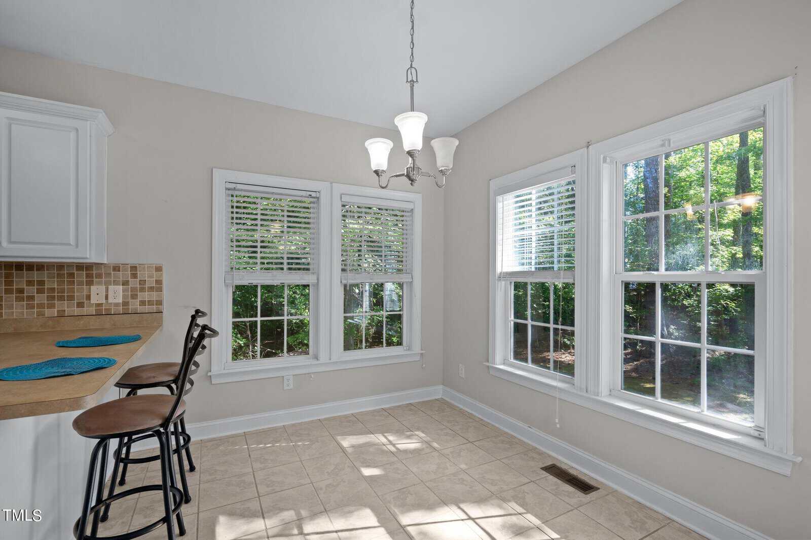 Bright breakfast nook with large windows, tile floor, and light fixture. Two barstools sit at a kitchen counter with blue mats. Sunlight streams in, and trees are visible outside through the windows.