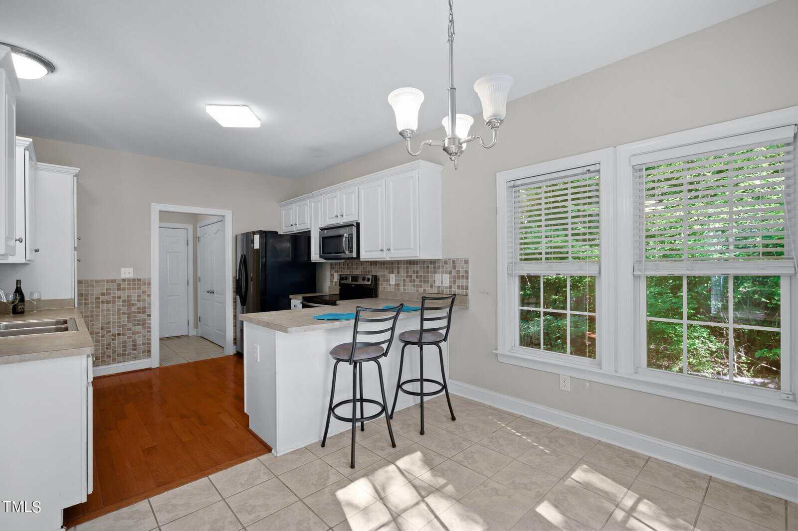 Bright kitchen with white cabinets, stainless steel appliances, a black refrigerator, a small island with two bar stools, tile flooring, hardwood accents, and large windows with white blinds overlooking greenery.