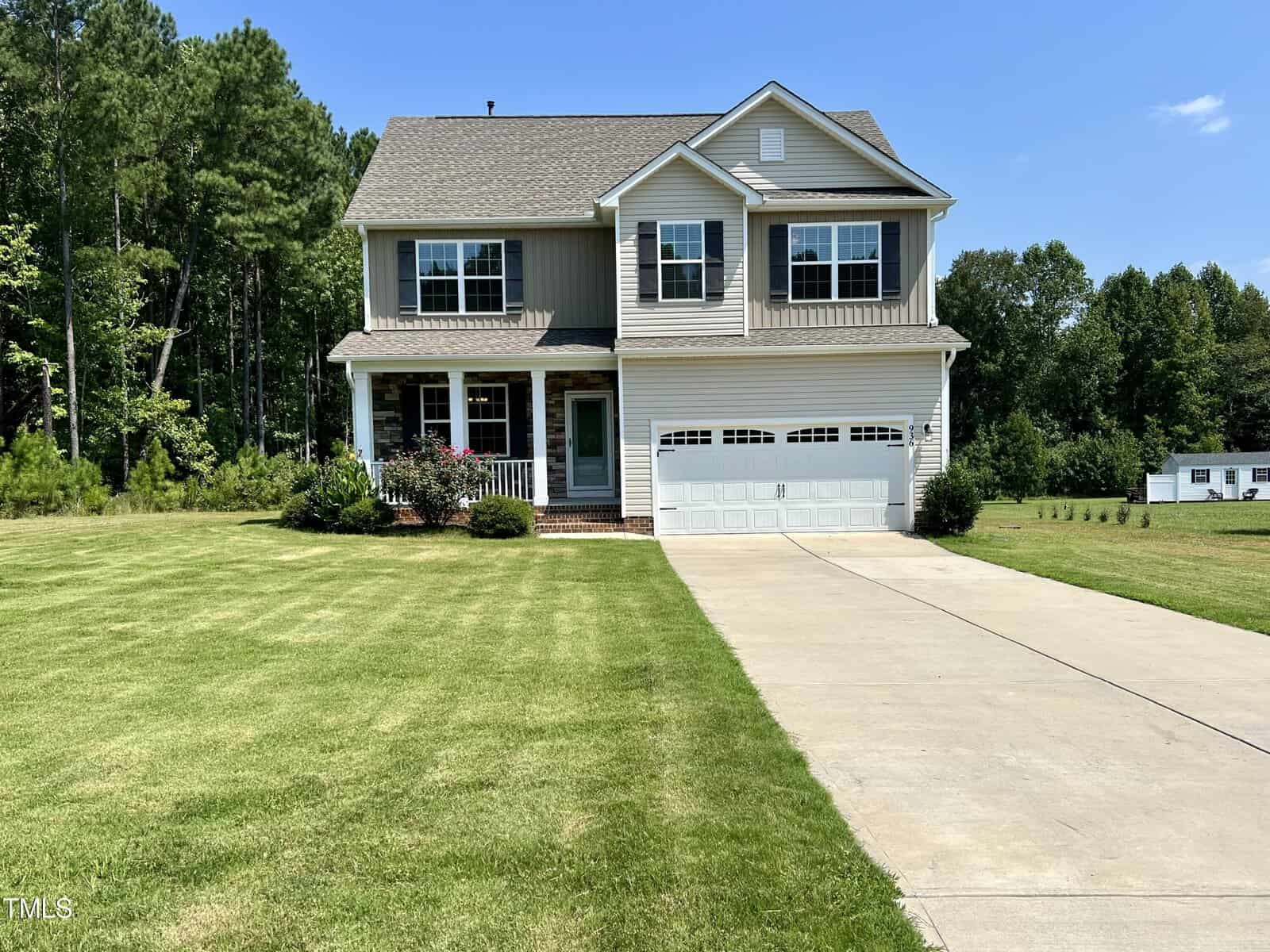 A two-story house with gray siding and a two-car garage is pictured. The house has a well-maintained front lawn, a driveway, and is surrounded by trees. There is a blue sky in the background.