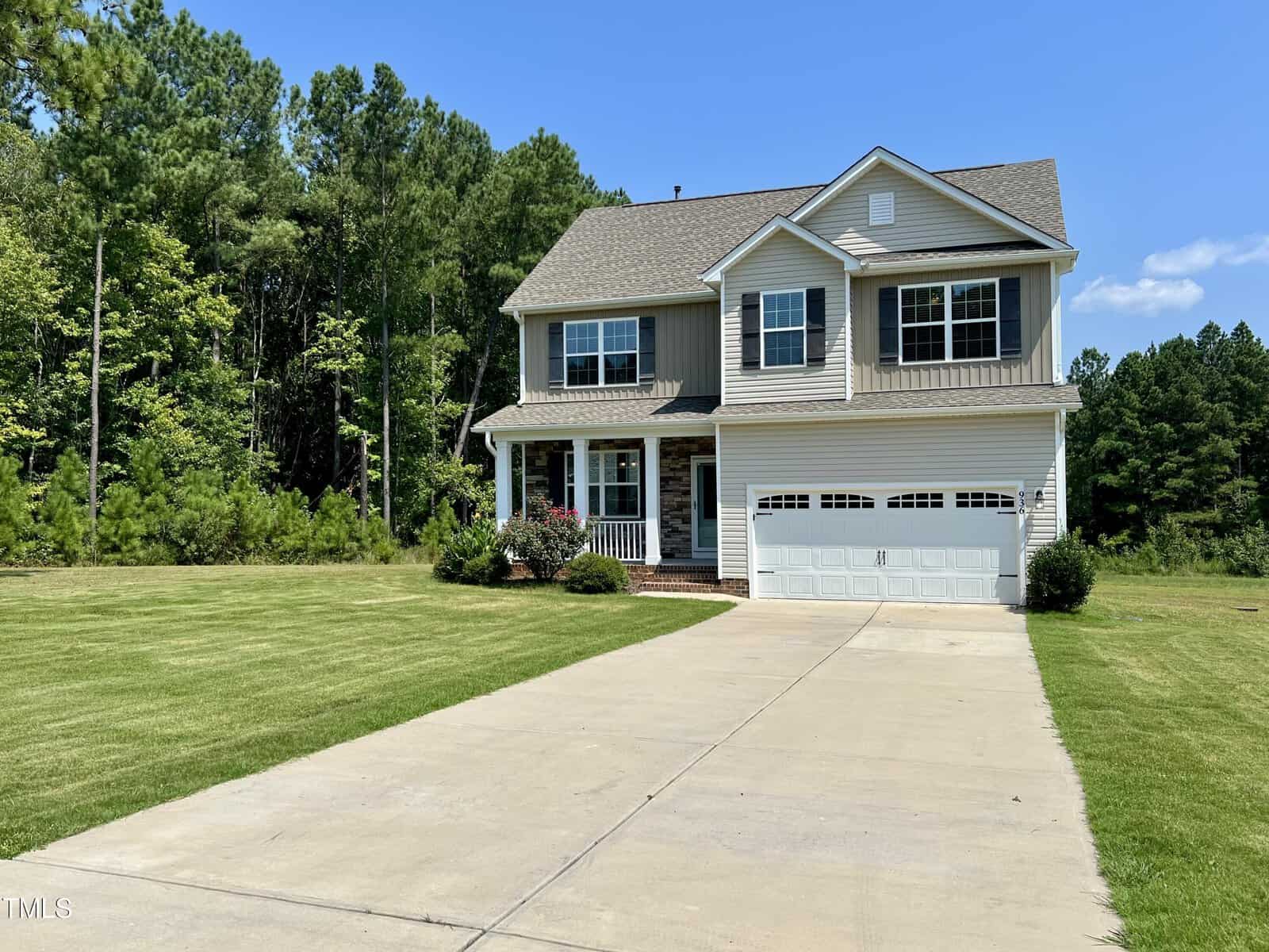 A two-story house with beige siding and white trim is situated in a lush green yard. It has a front porch, a two-car garage, and a long driveway. Trees line the background under a clear blue sky.