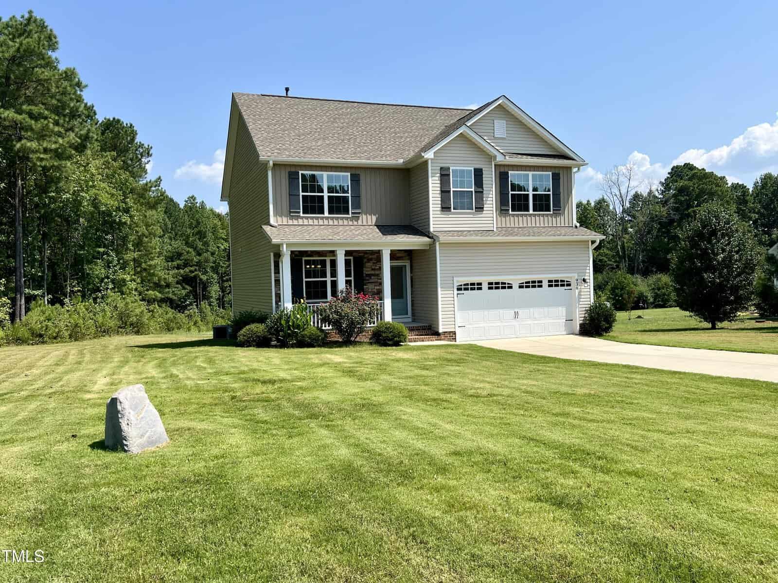 A two-story house with beige siding and a gabled roof is surrounded by a well-maintained lawn. A concrete driveway leads to a double garage. Trees frame the lush green yard under a clear blue sky. A small rock is in the front yard.