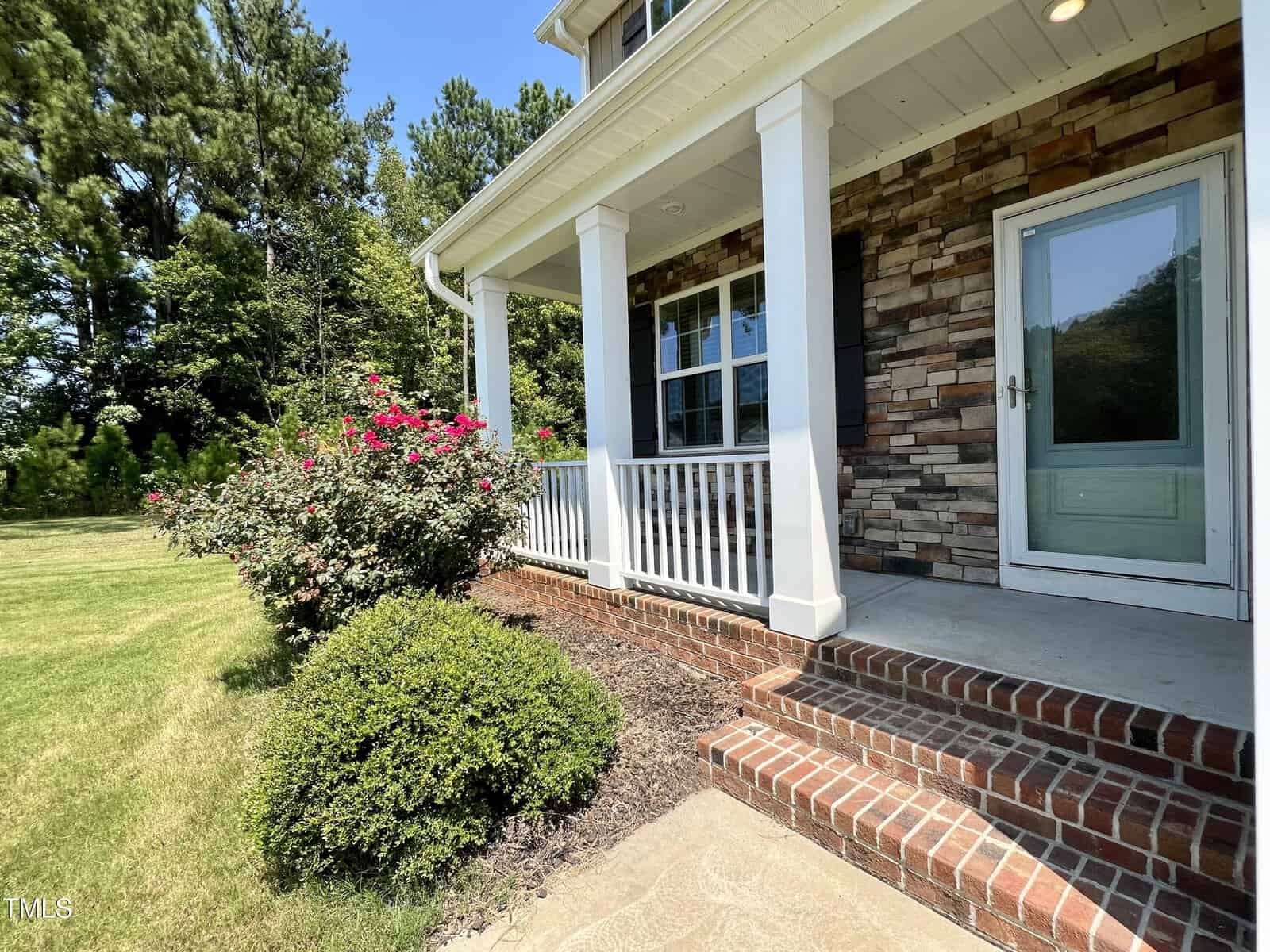 A house with a brick and stone facade features a small porch with white columns and railings. Red flowers and green shrubs line the walkway. A grassy lawn and trees are visible in the background under a clear blue sky.