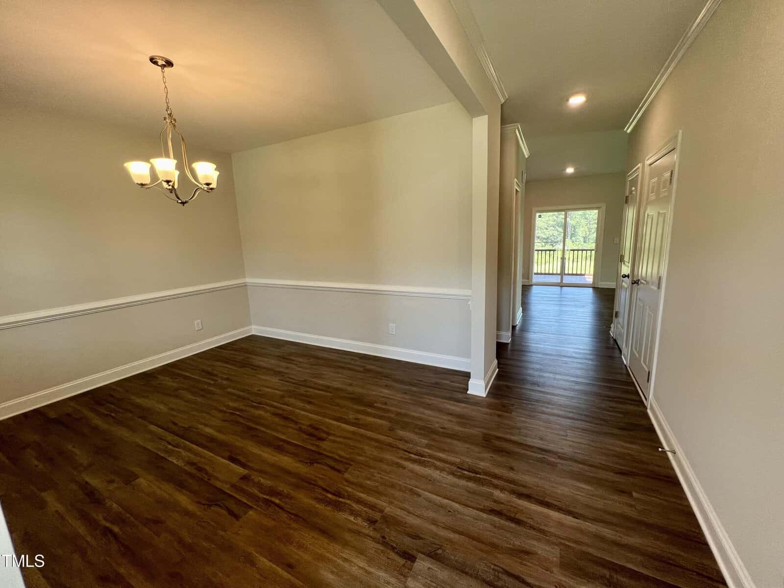 A room with wooden floors features an empty dining area with a chandelier, adjacent to a hallway. The hallway leads to double doors opening to a bright outdoor space. The walls are light-colored, and there is a white chair rail.