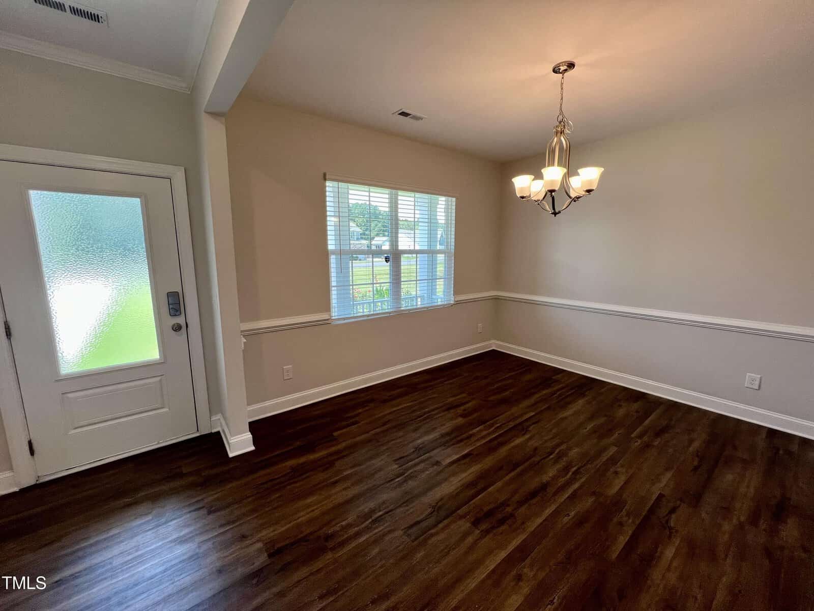 A room with hardwood flooring, white walls, and a decorative chair rail. A window with blinds is on one wall, and a frosted glass door is nearby. A chandelier with multiple lights hangs from the ceiling.