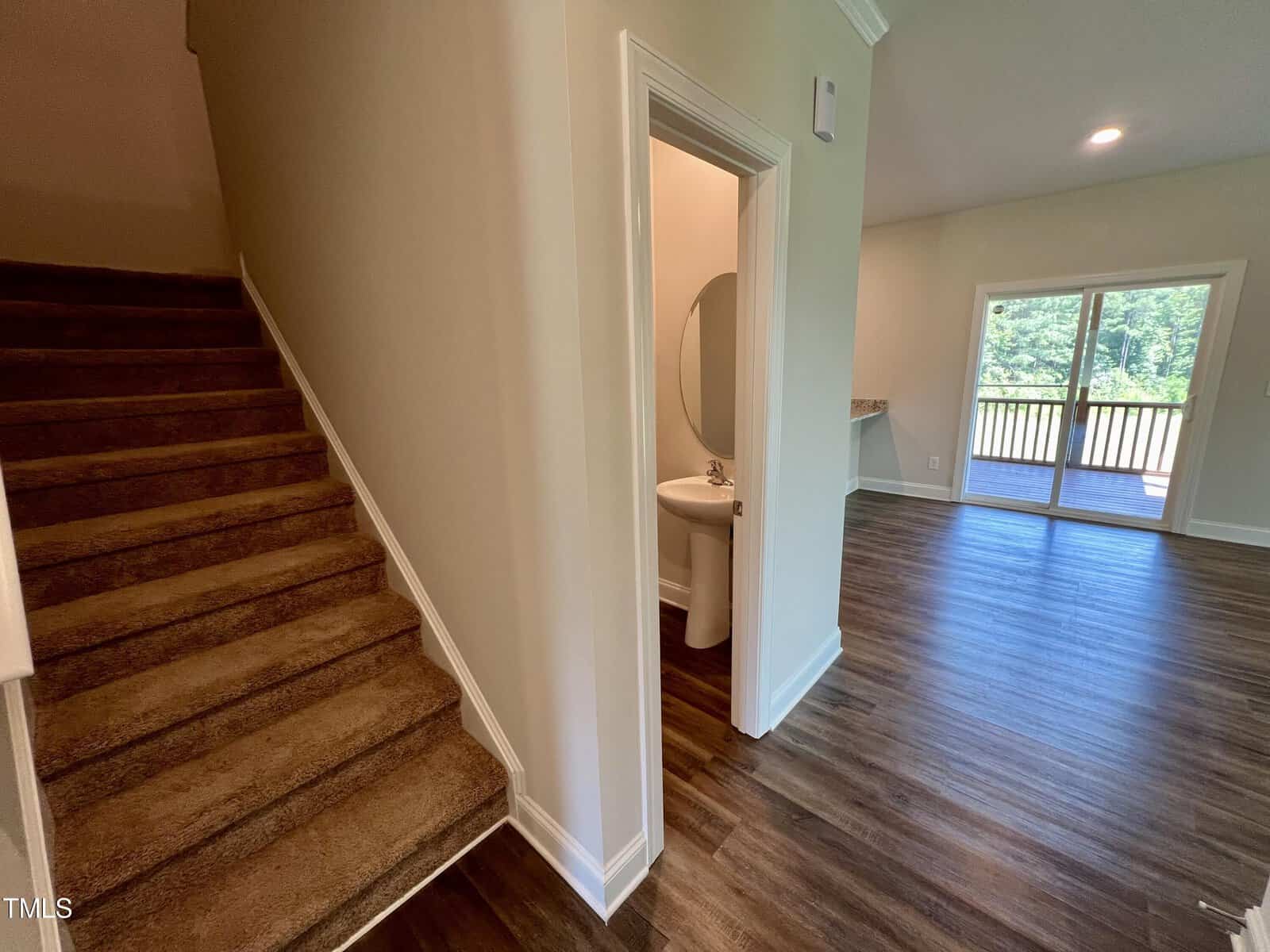 Interior view of a home showing a carpeted staircase on the left, a small bathroom with a pedestal sink in the middle, and a living area with wooden flooring leading to glass sliding doors opening to a balcony with a forest view on the right.